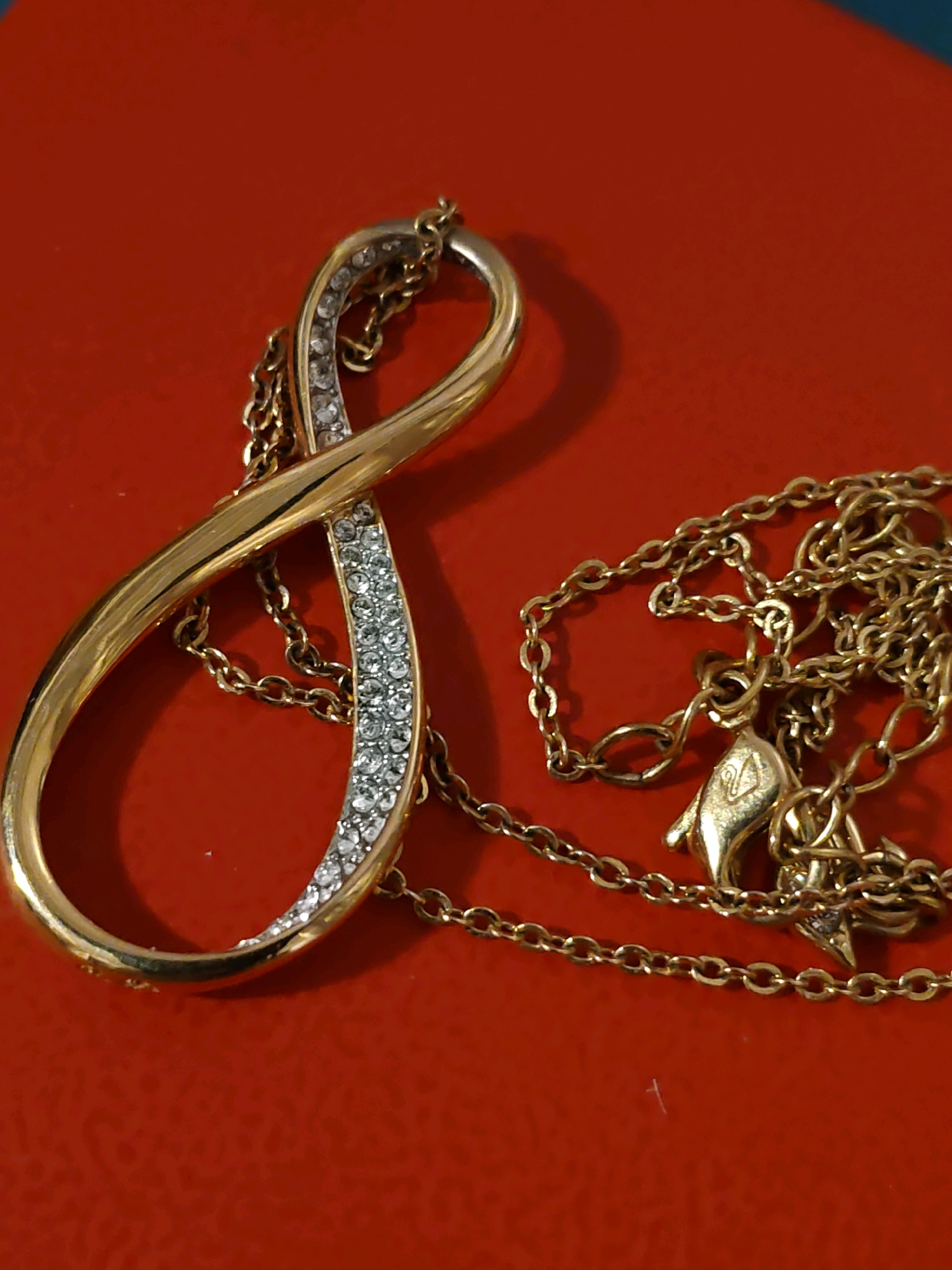 A gold pendant shaped like an infinity symbol, encrusted with small clear stones, is displayed on a vibrant red surface. A delicate gold chain is attached to the pendant and is draped around it. The scene is well-lit, with a slight shadow cast by the pendant on the left side. The background is a plain red surface. The image appears to be a product shot or a personal photograph of jewelry. There is no discernible information that places this image in Rostov-on-Don, Russia.