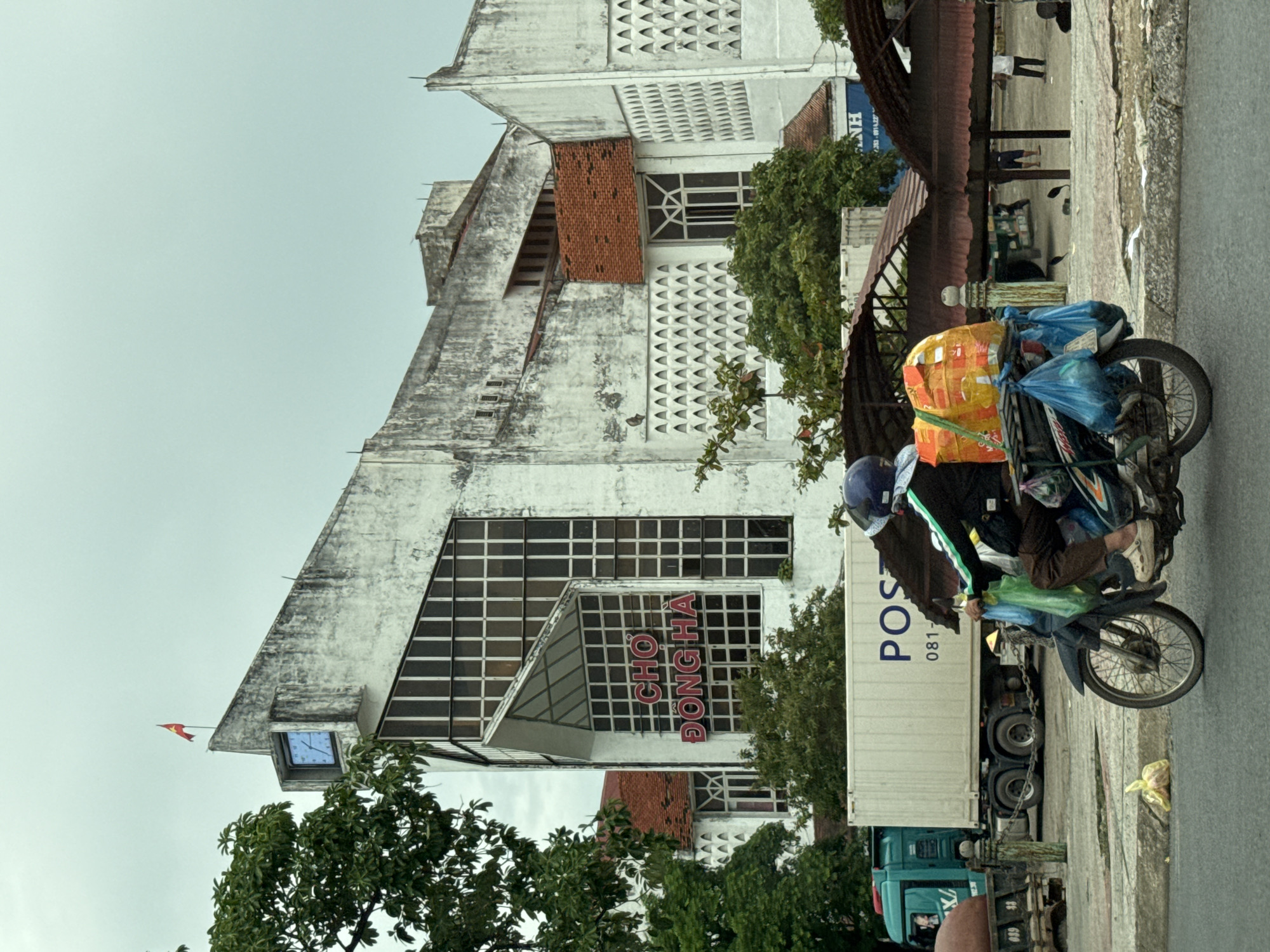 A person rides a motorcycle loaded with goods in front of a building with Vietnamese writing. The building is white and has a distinctive angular design. A large clock is visible on the upper left side of the building, and a flag is planted on the peak. The writing on the building reads "CHỢ ĐÔNG HÀ", which translates to Dong Ha Market. A large truck with "POST" written on its side is parked behind the motorcycle. The ground is paved, and there are some trees and awnings visible in the background. The sky is overcast.