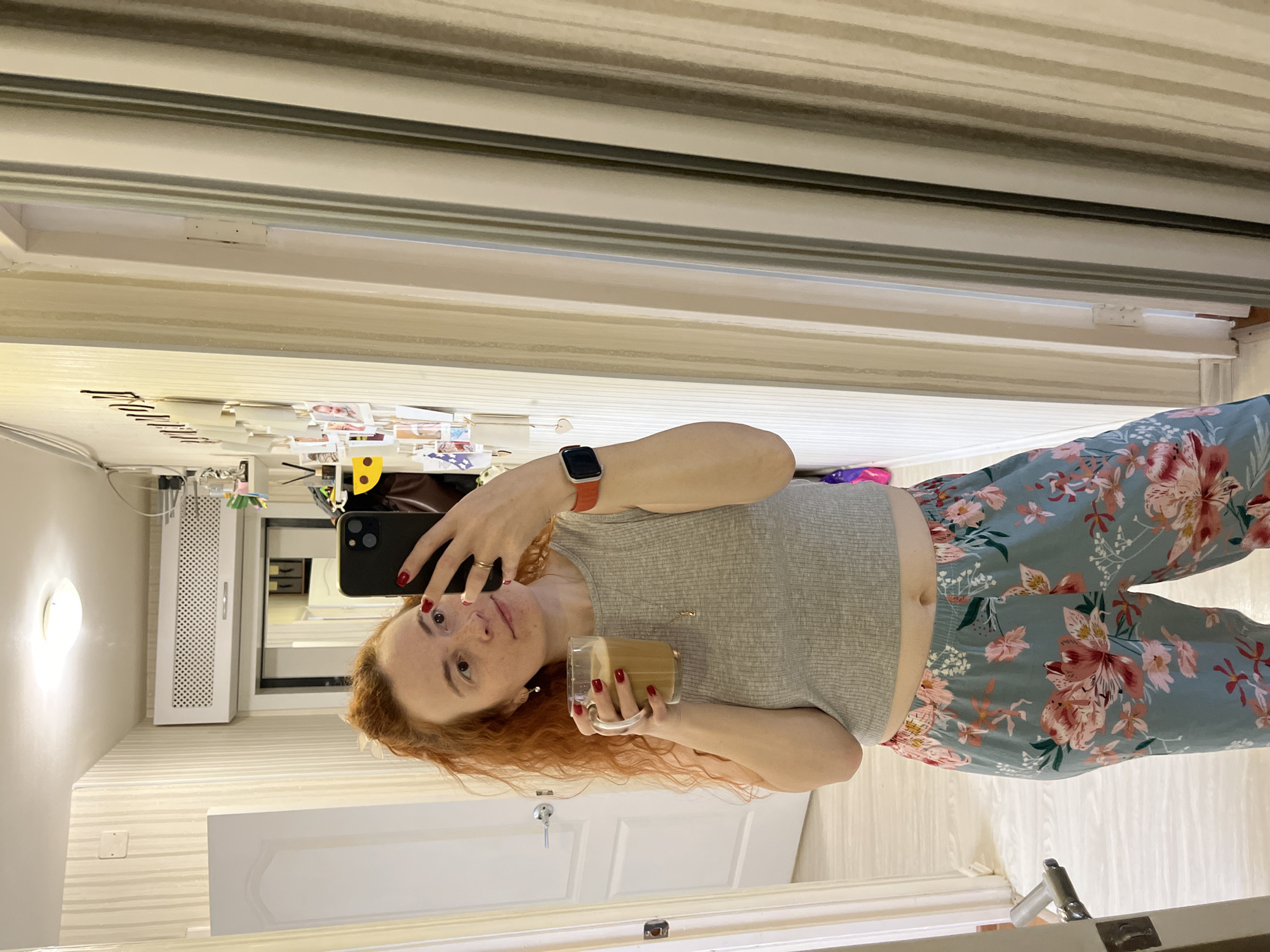 A red-haired, light-skinned person takes a selfie in a mirror, holding a glass of coffee. They are wearing a grey top and floral patterned pants. A smartphone is held in their left hand. The environment appears to be a residential interior, with a door, air conditioner, and various items on a shelf visible in the background. The location is Chaikovsky, Russia.