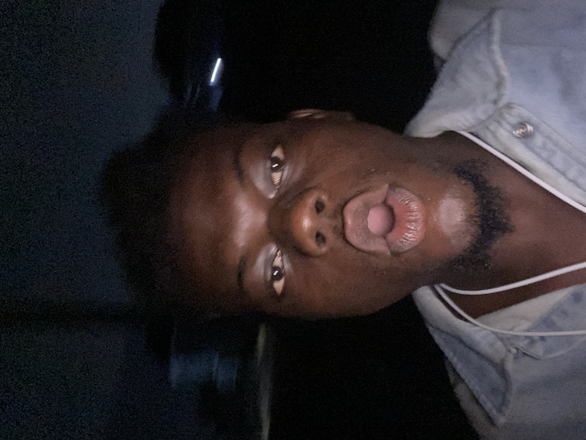 A man with dark skin is positioned in the center of the frame. He has short black hair, brown eyes, and thick lips parted in an "o" shape. He appears to be wearing a light blue denim jacket over a white collared shirt. The background is dark and indistinct, with a faint light source appearing at the top.