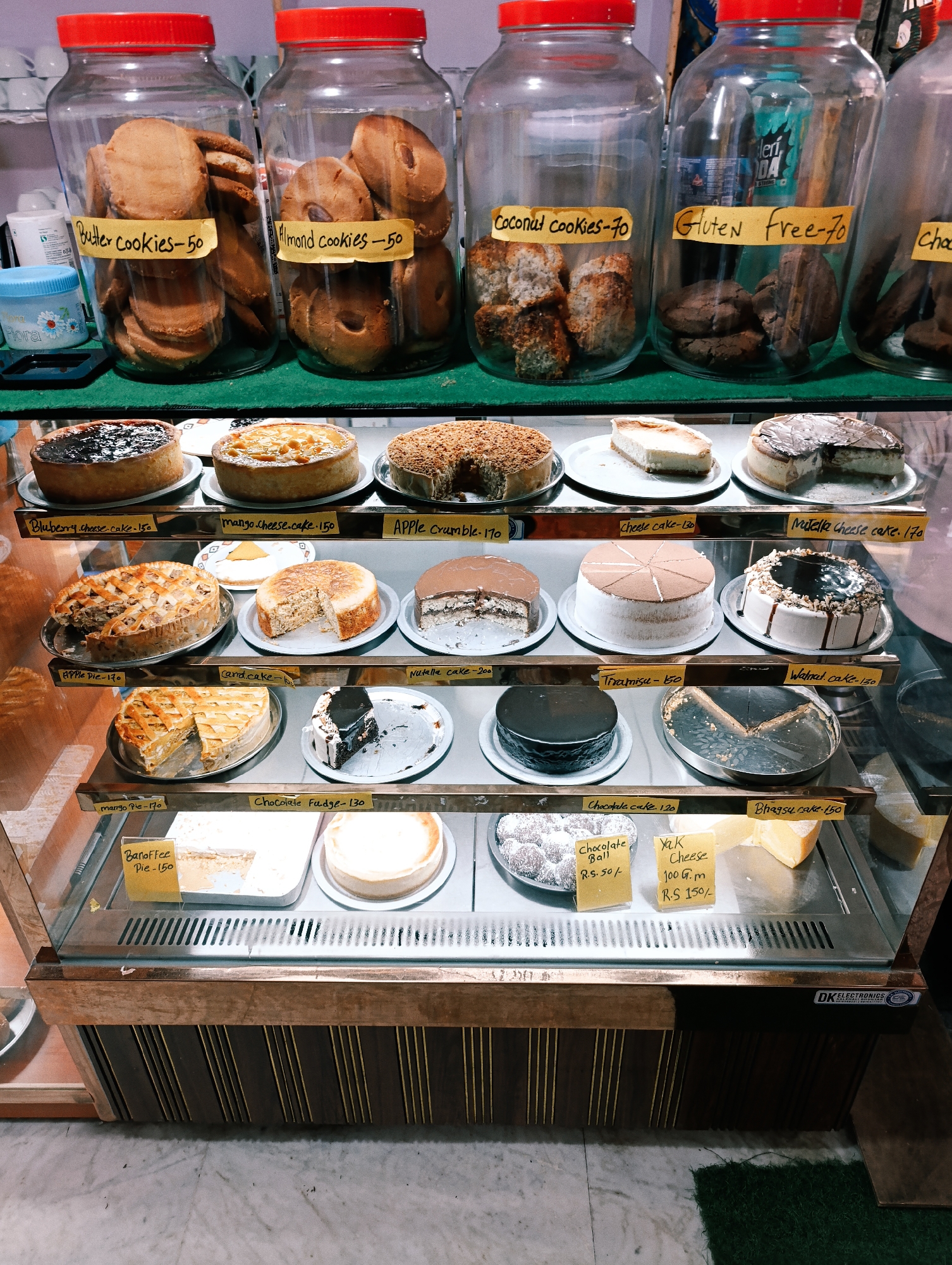 A glass-fronted display case filled with baked goods is presented at a bakery or cafe in Vashisht, India. The bakery is lit from above, creating a warm, inviting atmosphere. The display case is divided into multiple shelves. The top shelf holds four glass jars filled with cookies. From left to right, the labels read "Butter cookies -50," "Almond cookies -50," "Coconut cookies -70," and "Gluten Free -70." Below the jars, the first shelf of the display case holds four whole cakes. From left to right, these are a blueberry cheesecake, a mango cheesecake, an apple crumble cake, and a cheesecake. The second shelf displays four items: an apple pie, a round cake, a chocolate-covered cake, and another round cake with chocolate frosting. The third shelf features a chocolate cake, a slice of chocolate cake with white icing, and a round cake with chocolate frosting and nuts. The bottom shelf holds a chocolate cake, a slice of cake, and two pieces of cheese, one labeled "Yak Cheese 100 G.m R.S 150/-". The background shows a wall with wooden paneling and a glimpse of a tiled floor.