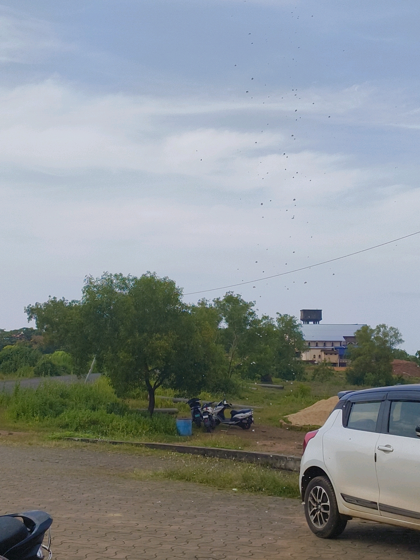 The image captures a street scene in Bhatkal, India, on a cloudy day. In the foreground, a white car with a black roof is parked on a patterned pavement. To the left of the car, part of a black motorcycle is visible. Behind the vehicles, a patch of green grass and vegetation leads to a larger tree with dense foliage. Further in the background, several motorcycles are parked, and a blue bin is present. Beyond the greenery, a sand pile is visible, and a building with a water tank on top can be seen in the distance. The sky is overcast with scattered clouds, and numerous small dark specks, possibly insects or dust, are visible throughout the frame, particularly in the upper portion of the image.