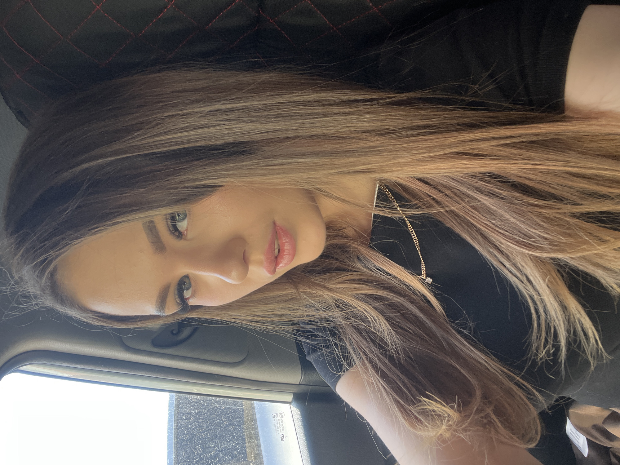A woman with long, light brown hair is sitting in a car. She is wearing a black shirt and a gold necklace. The interior of the car is dark, with a window visible on the left side. The background outside the window is blurry. The location is not identifiable as Chaikovsky, Russia, from the visual information provided.