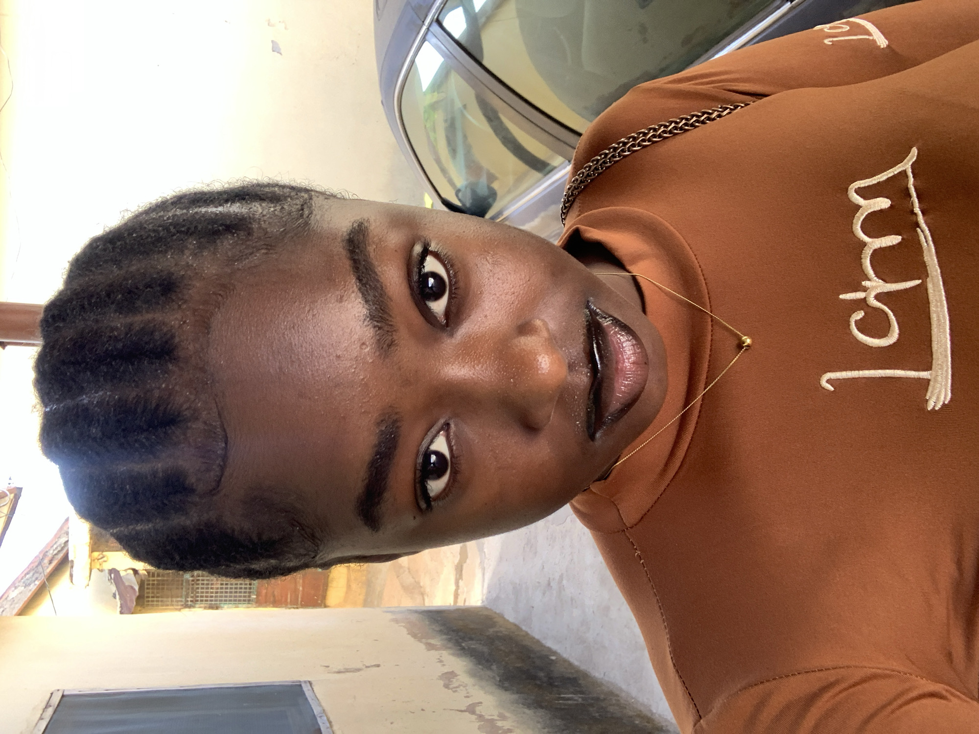 A medium shot depicts a Black female in Rafin Par, Nigeria. She is wearing a brown top with a small gold chain. Her hair is styled in braids. A car is partially visible in the background. The setting appears to be near a building exterior.