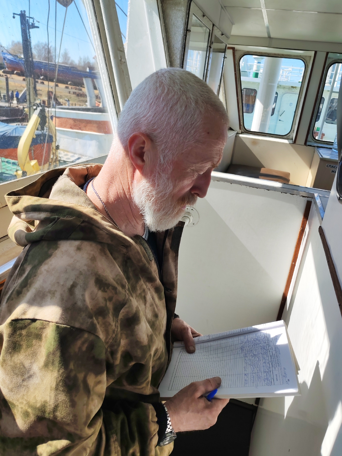 A man with white hair and a white beard is the central subject, positioned in the foreground and looking down at a notebook he holds. He is wearing a camouflage-patterned jacket. He appears to be focused, possibly reading or writing in the notebook, which has ruled lines and handwritten entries. He holds a blue pen in his right hand, and a watch with a metal band is visible on his left wrist.
The man is inside what appears to be the bridge or cabin of a vessel, with large windows surrounding him. The interior is functional, featuring light-colored walls and some wooden trim. Bright sunlight streams in through the windows, casting shadows and highlighting the man and the notebook.
Through the windows, a bustling outdoor scene is visible, strongly suggesting a shipyard or boat maintenance area. In the background, there are several boats or ships on land, some appearing to be in various stages of repair or construction, including a prominent blue-hulled vessel. A crane and other structures are also visible. The clear sky indicates good weather conditions.
On a white panel to the man's right, partially visible text in what appears to be Cyrillic script reads "СПАСА" and "ЖИ", likely part of a safety sign such as "Спасательный жилет" (Life vest). The notebook itself contains multiple lines of handwritten text and numbers, suggesting it is a logbook or record of some kind.