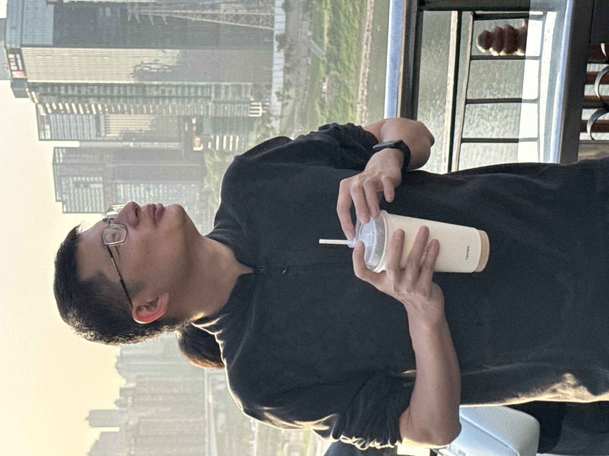 A man with glasses stands on a balcony, holding a cup with a straw, possibly a beverage, in his hands. The background features a cityscape, including visible high-rise buildings. The location appears to be the Yuzhong District, China, judging by the architectural style and urban environment in the background. The man's action is simply holding the cup. He is also wearing a dark-colored short sleeved shirt, and a black watch on his wrist.