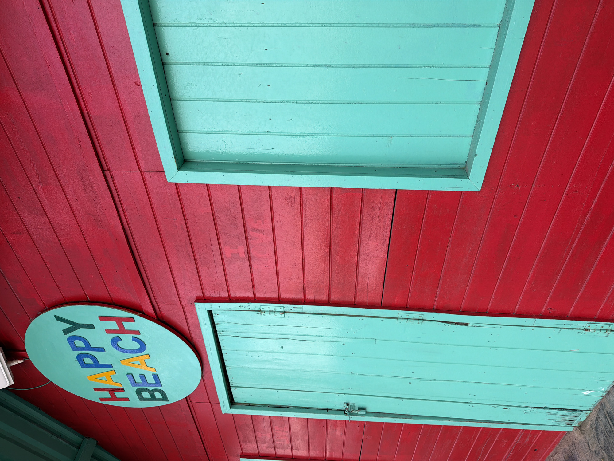 A bright red wooden building with teal trim is shown in Nha Trang Ward, Vietnam. A round teal sign with colorful block letters spells out "HAPPY BEACH" and is mounted on the building's exterior. To the left of the sign is a teal wooden door, and to the right is a teal wooden window. The building's red wooden planks run horizontally. The image is a close-up, focusing on the architectural details and signage of the building. No people are visible in the image.