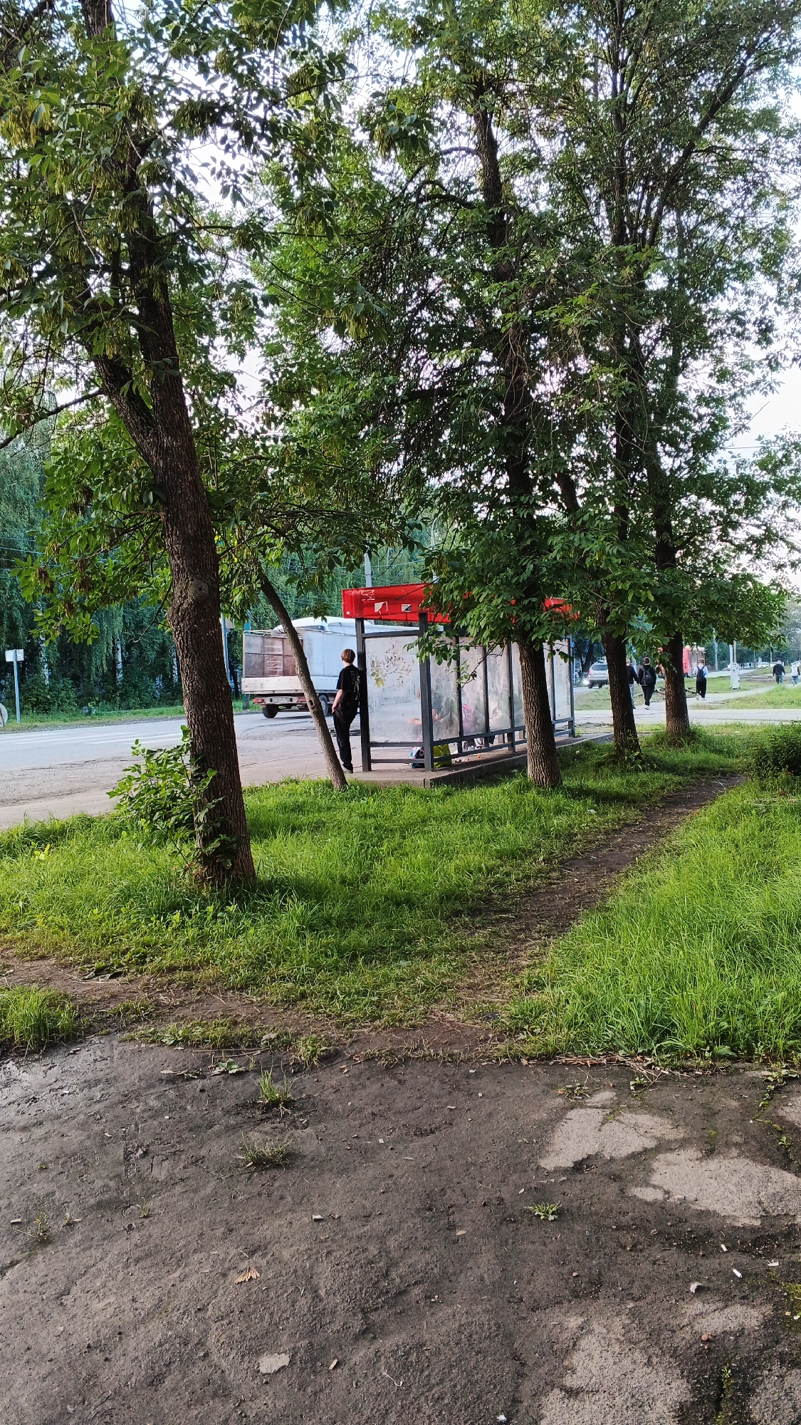 A medium shot shows a bus stop shelter on a cloudy day in Izhevsk, Russia. A young person in a black t-shirt and pants stands at the entrance of the shelter, facing away from the camera. The shelter has a red roof and glass panels. Trees with green leaves are positioned in front of and to the side of the shelter, with a dirt path leading away from it through lush green grass. A road with a white truck and other vehicles is visible in the background, along with trees and buildings. The foreground consists of a dirt and asphalt surface.