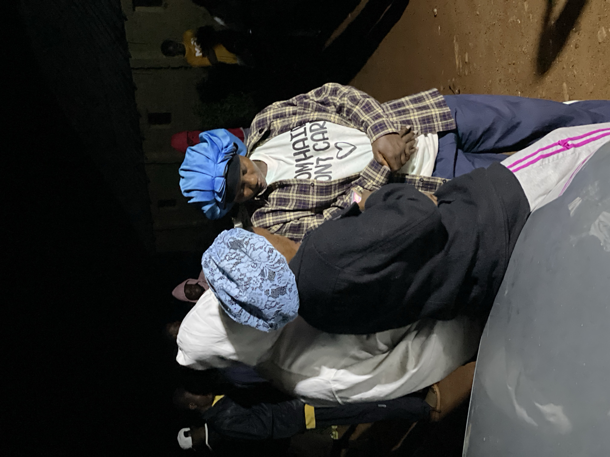 In Wumat, Nigeria, at night, a group of individuals are gathered outdoors near a building. Two women are in the foreground, both wearing bonnets. The woman on the right, wearing a blue bonnet, is dressed in a plaid shirt over a white t-shirt with the text "I DON'T CARE" and a heart symbol. She is looking down. The woman on the left, wearing a light blue lace bonnet, faces away from the camera and is dressed in a black jacket. A person in a white hooded garment is positioned between them. Other individuals are visible in the background, including one in a yellow shirt. The ground appears to be dirt.