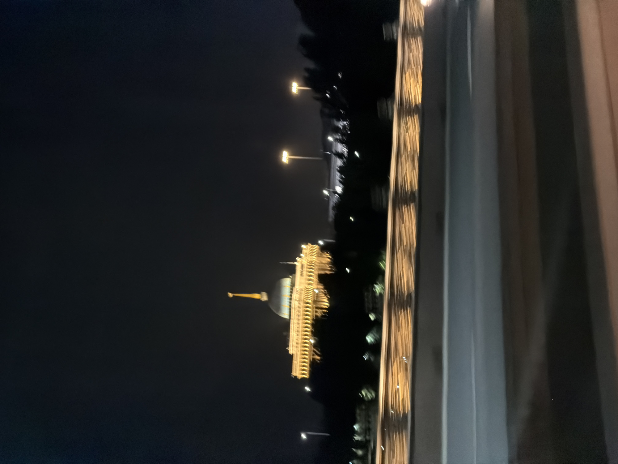 The image, taken at night, shows a large, illuminated building in Astana, Kazakhstan. The building has a prominent dome topped with a spire. Its facade is decorated with numerous lights, giving it a grand appearance. In the foreground, a road or highway is visible, with a railing along its edge that is also lit with lights. The sky is dark, indicating nighttime. The overall scene suggests an urban environment with significant architecture and infrastructure illuminated for nighttime viewing.