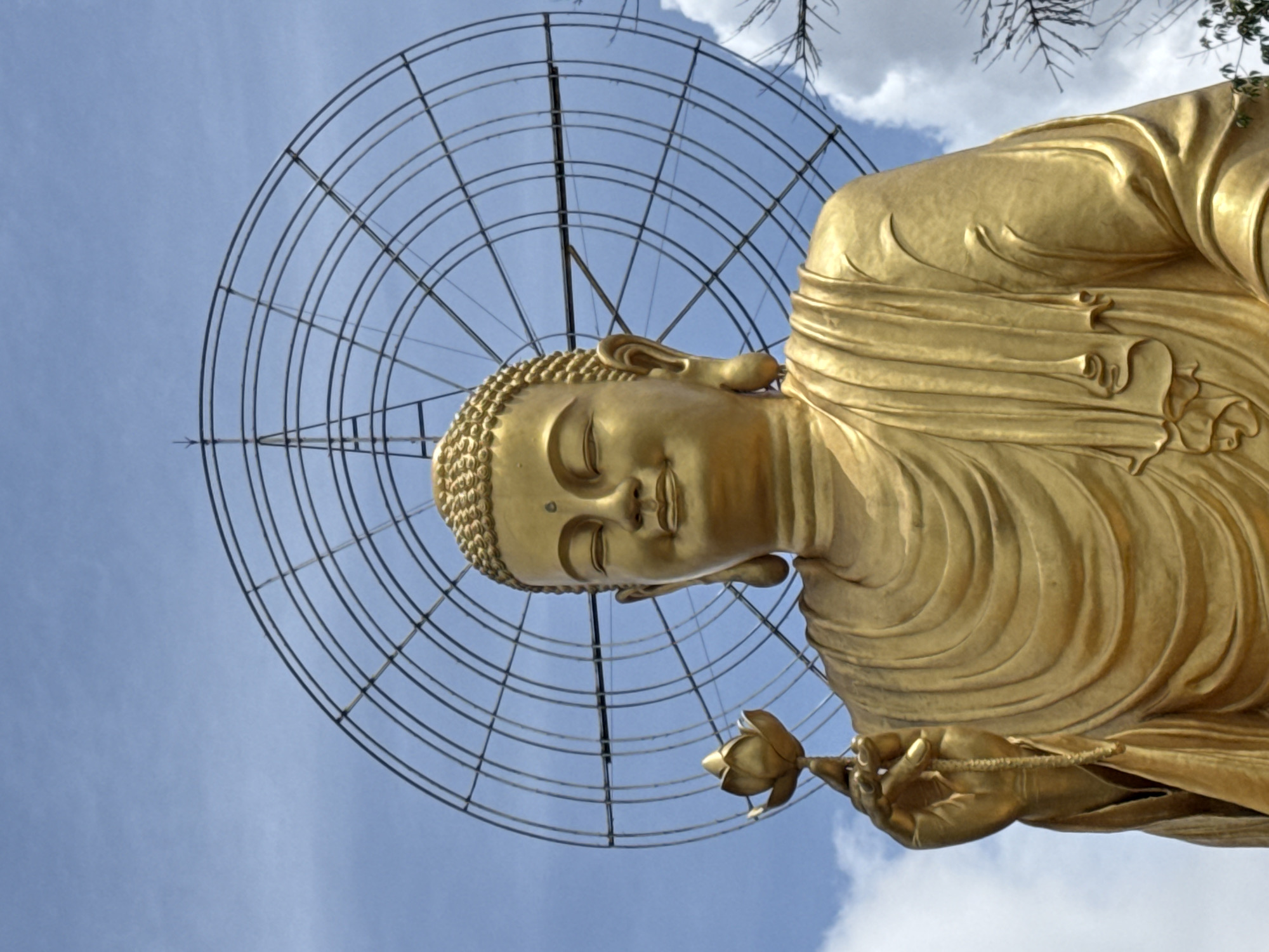 A close-up, low-angle shot captures a golden Buddha statue holding a lotus flower against a backdrop of a bright blue sky with scattered white clouds. Behind the statue, a large, circular metal structure with radiating spokes is visible. The Buddha's eyes are closed, and a gentle smile graces its face. The statue is draped in traditional robes, with intricate folds and creases. The image appears to be taken outdoors, with the lighting suggesting it is daytime. The location is specified as Lâm Viên - Đà Lạt, Vietnam.