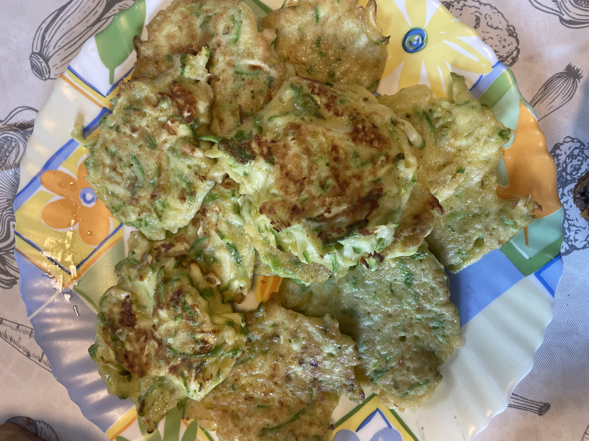 The image shows a plate of zucchini fritters on a patterned tablecloth. The fritters are golden brown and appear to be made with shredded zucchini and batter. They are arranged in a pile on a colorful, scalloped-edge plate with a floral design. The tablecloth has a greyish-white background with black line drawings of vegetables. The scene is likely indoors, possibly in a home kitchen. There are no people or animals visible in the image. The location is not specified in the image, and therefore, it cannot be confirmed to be in the city of Турово, Russia.