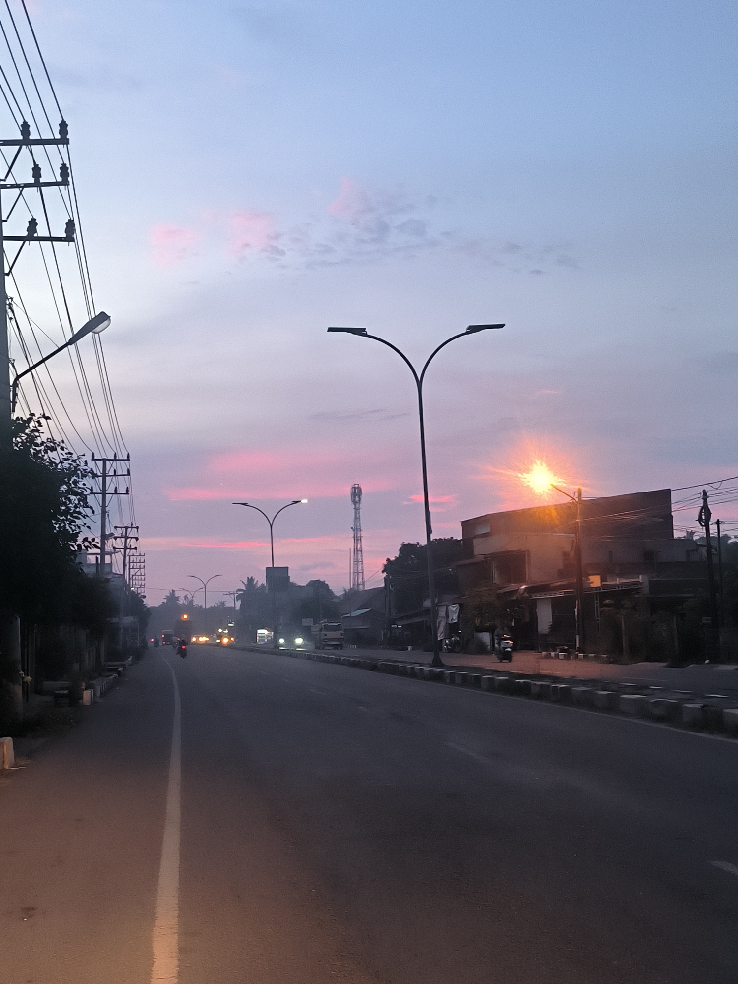 The image depicts a street scene in Lhokseumawe, Indonesia, at dusk or dawn. The sky is a gradient of pale blue and pink, with wisps of clouds tinged with color. A main road dominates the foreground, with a white line marking the center. On the left side of the road, a sidewalk is visible with several white concrete bollards. Tall utility poles with numerous wires run along this side, along with a dense line of dark trees.
Streetlights, some with a distinctive double-arm design, are present on both sides of the road. On the right side, buildings with shops or residences line the street, some appearing to be two stories high. A prominent communication tower is visible in the distance. Several vehicles, including motorcycles and what appear to be cars and trucks, are present on the road, with their headlights and taillights illuminated. A few individuals are seen on motorcycles, suggesting light traffic. The overall impression is a typical urban or semi-urban street in Indonesia during the transitional light of day and night.