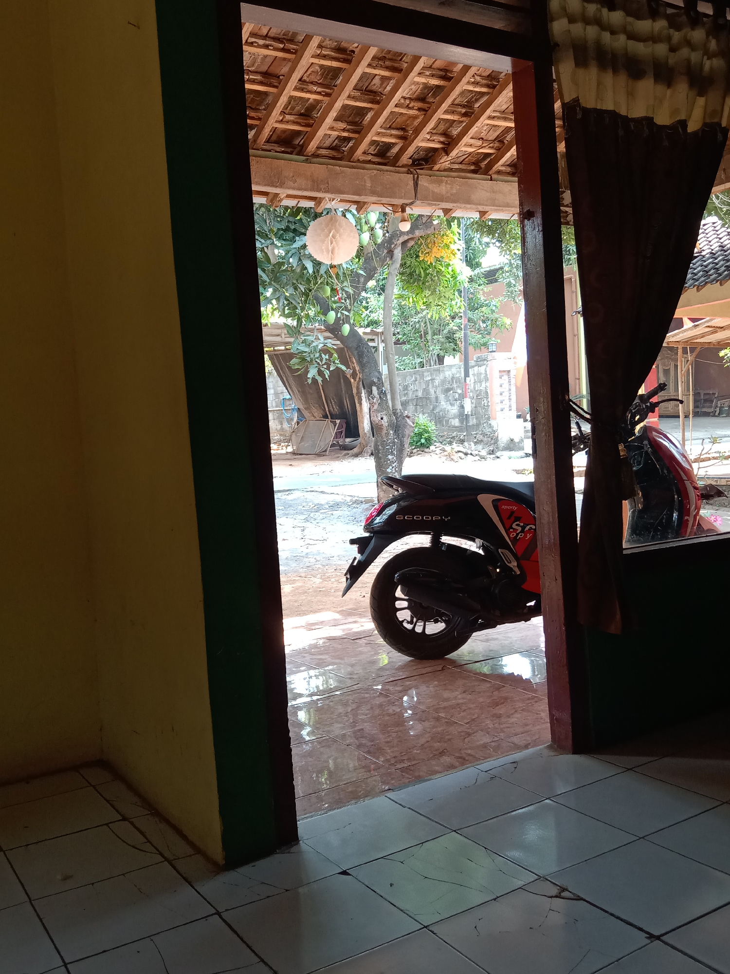 The image captures an outdoor scene viewed from an interior doorway in what appears to be a residential setting in Menganti, Indonesia. The foreground shows a tiled floor, with a yellow wall and a dark green doorframe on the left. Through the open doorway, a street is visible with a red and black scooter parked on the right side. The scooter has the word "SCOOPY" written on its side. Beyond the scooter, trees with hanging fruit, a brick wall, and various structures are present, suggesting a somewhat rural or suburban environment. The roof of the house is partially visible, constructed with clay tiles. A decorative white orb hangs from a tree branch. The overall impression is a typical Indonesian dwelling with its surroundings. There are no people visible in the image.