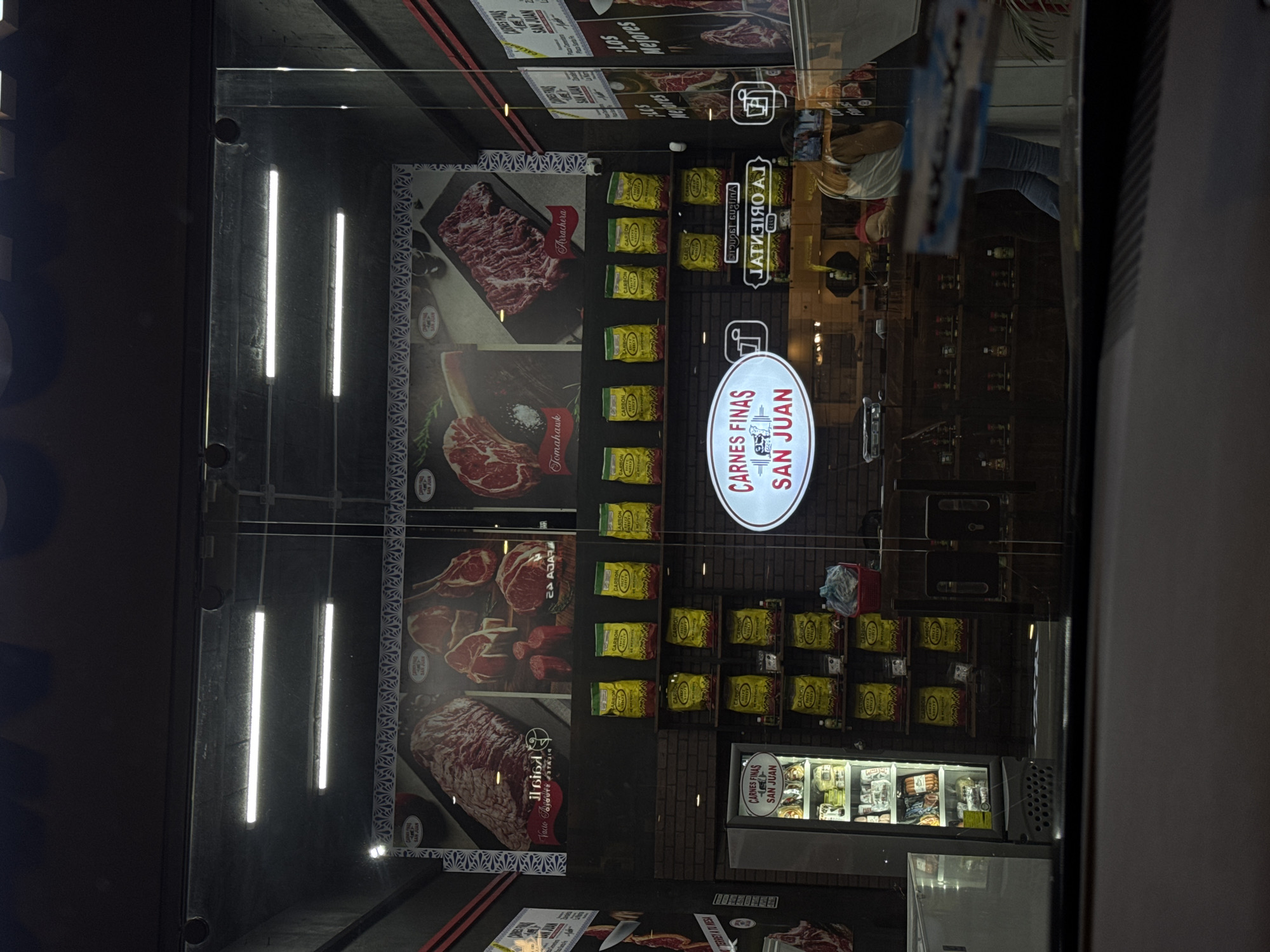 A display window with the words "CARNES FINAS SAN JUAN" is seen. Posters depicting various cuts of meat, such as "Arrachera", "Tomahawk", and "Vacio Tagurdes" are prominently displayed. Below are stacks of yellow bags, likely containing products related to the meat selection. In the reflection, a glimpse of the interior shows shelves stocked with various items, and a small refrigerated display case filled with food products. There is a partial reflection of a person in the window. The background and the reflections within suggest this is an urban setting, likely a store front in Puebla, Mexico, given the text on the signage.
