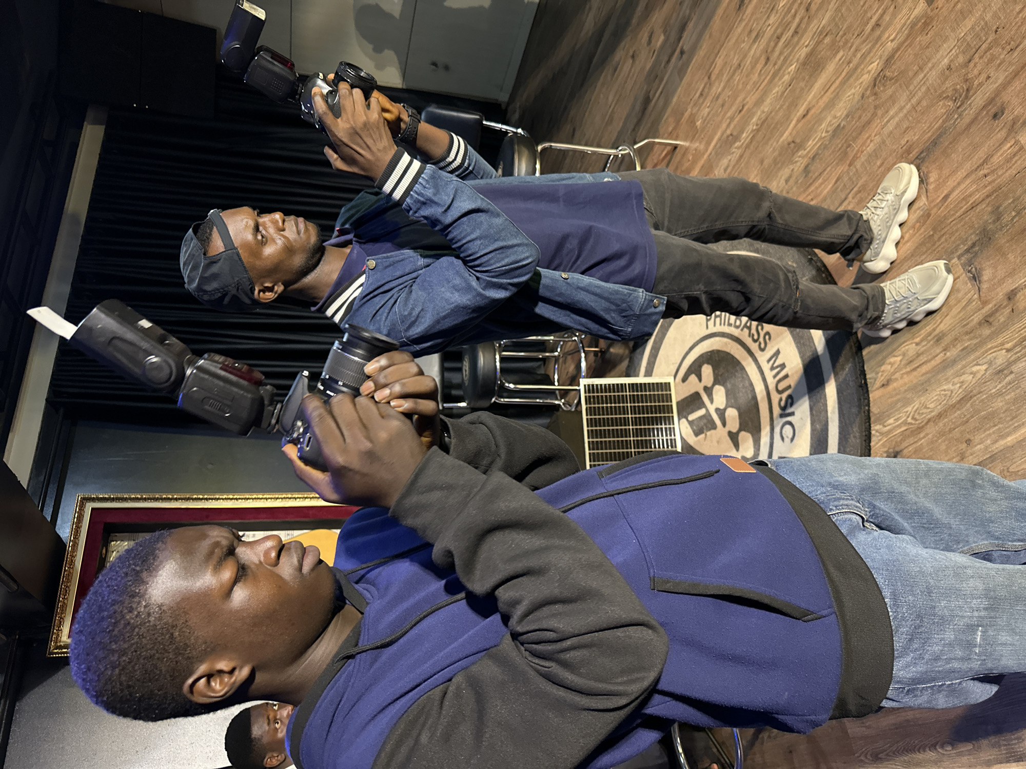 The image depicts a medium shot of two young men, likely photographers, in an indoor venue that appears to be a studio or performance space. The men are holding cameras with external flashes attached, suggesting they are actively documenting an event. They are both looking down at their cameras, focused on their subjects.
The man in the foreground, on the left, is wearing a blue hooded sweatshirt with black sleeves and a black drawstring. He is also wearing blue jeans. His camera is held with both hands, and his brow is furrowed in concentration.
The second man, on the right, is wearing a denim jacket over a dark blue t-shirt and dark pants. He has a black baseball cap on backward. His camera is also held with both hands.
The environment includes a wooden floor, a dark curtain in the background, and some stools. There is a framed artwork or photograph on the wall behind the man in the foreground. A circular rug with text that appears to be "PHILIDASS MUSIC" is visible on the floor between the two men. A small, rectangular grate or vent is also present on the floor.
The lighting suggests an indoor setting with artificial lights, as indicated by the flashes on the cameras. The overall impression is one of a professional or semi-professional photography setup. Without further context, it is impossible to determine the specific location within Barakin Lerawi, Nigeria.