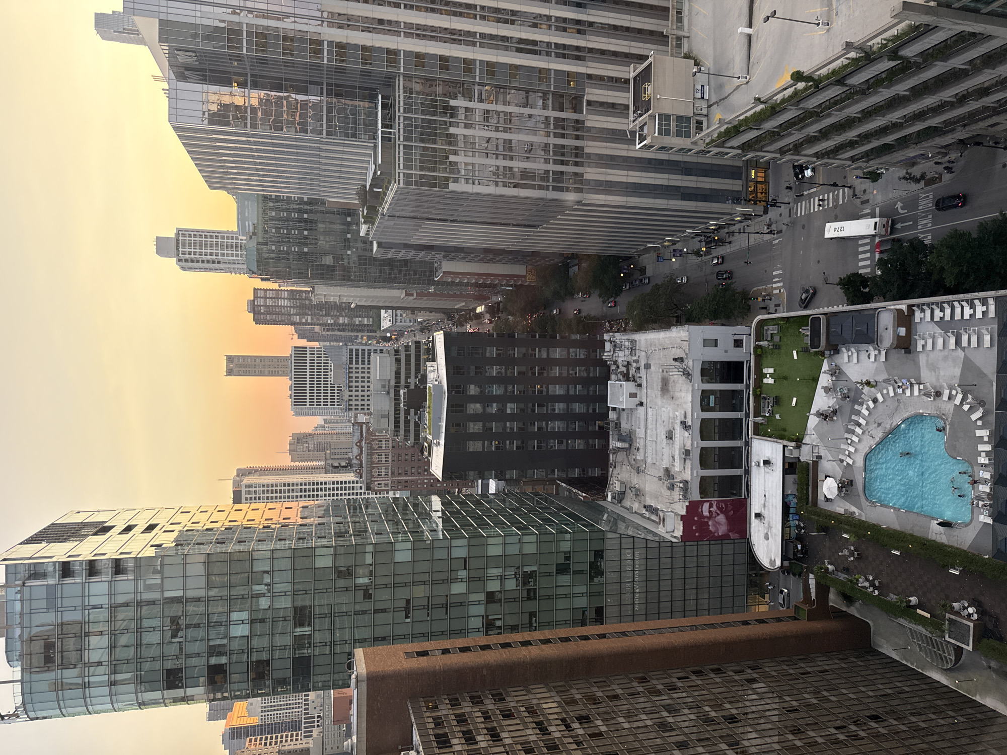 This is a high-angle, vertical, exterior, daytime, urban scene in Chicago, Illinois, USA. The image features a dense cityscape with numerous high-rise buildings of varying architectural styles. The sky is a warm gradient of orange and yellow, suggesting either sunrise or sunset. In the foreground, a rooftop swimming pool area with lounge chairs and umbrellas is visible. Several people are in the pool. Adjacent to the pool area, there are outdoor seating areas with tables and chairs, as well as some greenery. A street with moderate traffic, including cars and a bus, runs through the scene. Reflections of the sky and surrounding buildings are visible on the glass facades of many of the skyscrapers. The overall impression is one of a bustling urban environment captured from a considerable height.