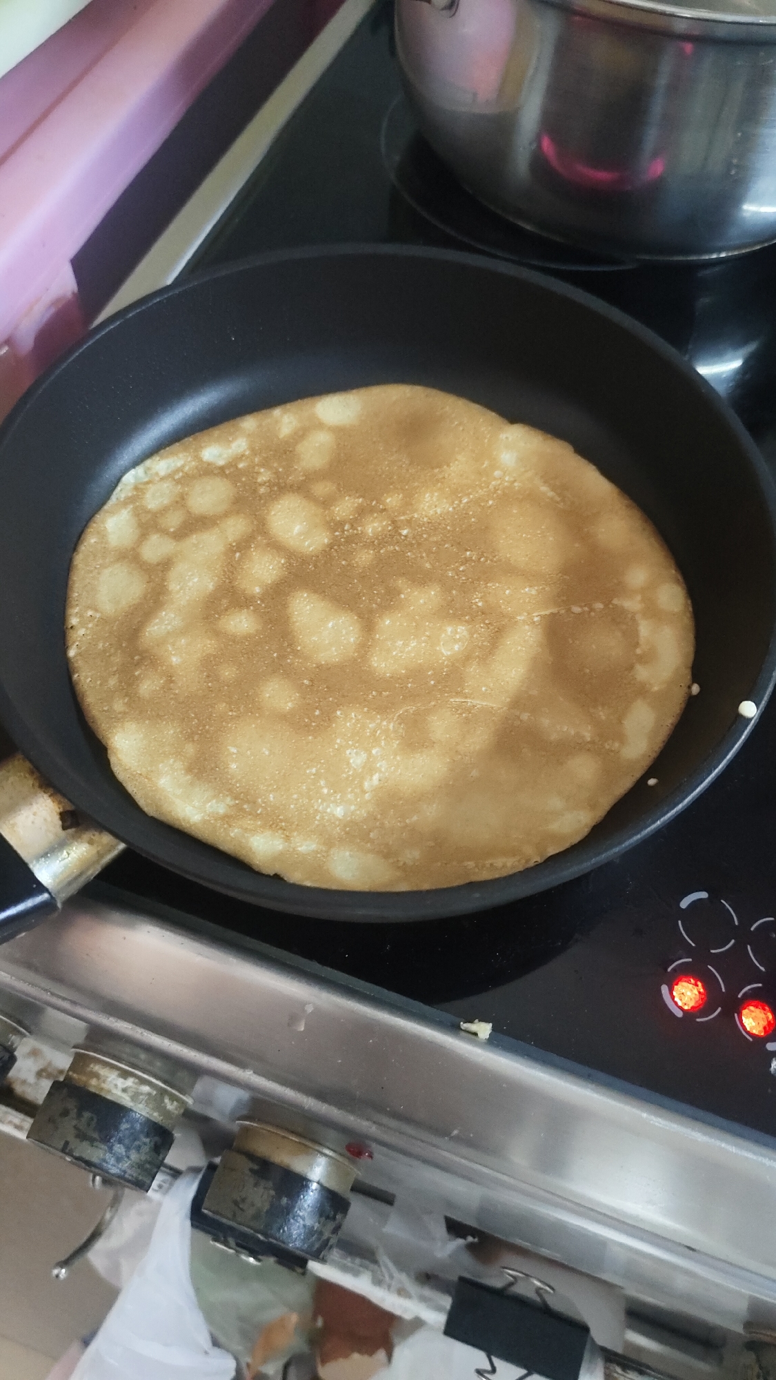 This image captures a domestic cooking scene in a kitchen in Krasnoyarsk, Russia, focusing on a pancake being prepared. The central subject is a round, golden-brown pancake, appearing lightly cooked and dotted with lighter, bubbled areas, filling most of a black non-stick frying pan.
The frying pan rests on a black ceramic or induction stovetop, which has red indicator lights illuminated on its control panel, signifying that a burner is active. To the upper right, a shiny, stainless steel pot is also visible, sitting on another section of the stovetop.
Below the cooking surface, the edge of a metallic kitchen counter or range separates the cooking area from the lower part of the scene. Underneath this edge, two older, dark-colored control knobs with silver tops are visible. Further down and to the left, there's some clutter including a white plastic bag and crumpled brown paper or possibly food waste. In the upper left background, a portion of a pink or purplish object, possibly a storage container or part of the kitchen wall, is visible. The lighting suggests an indoor setting. No discernible text is visible in the image.