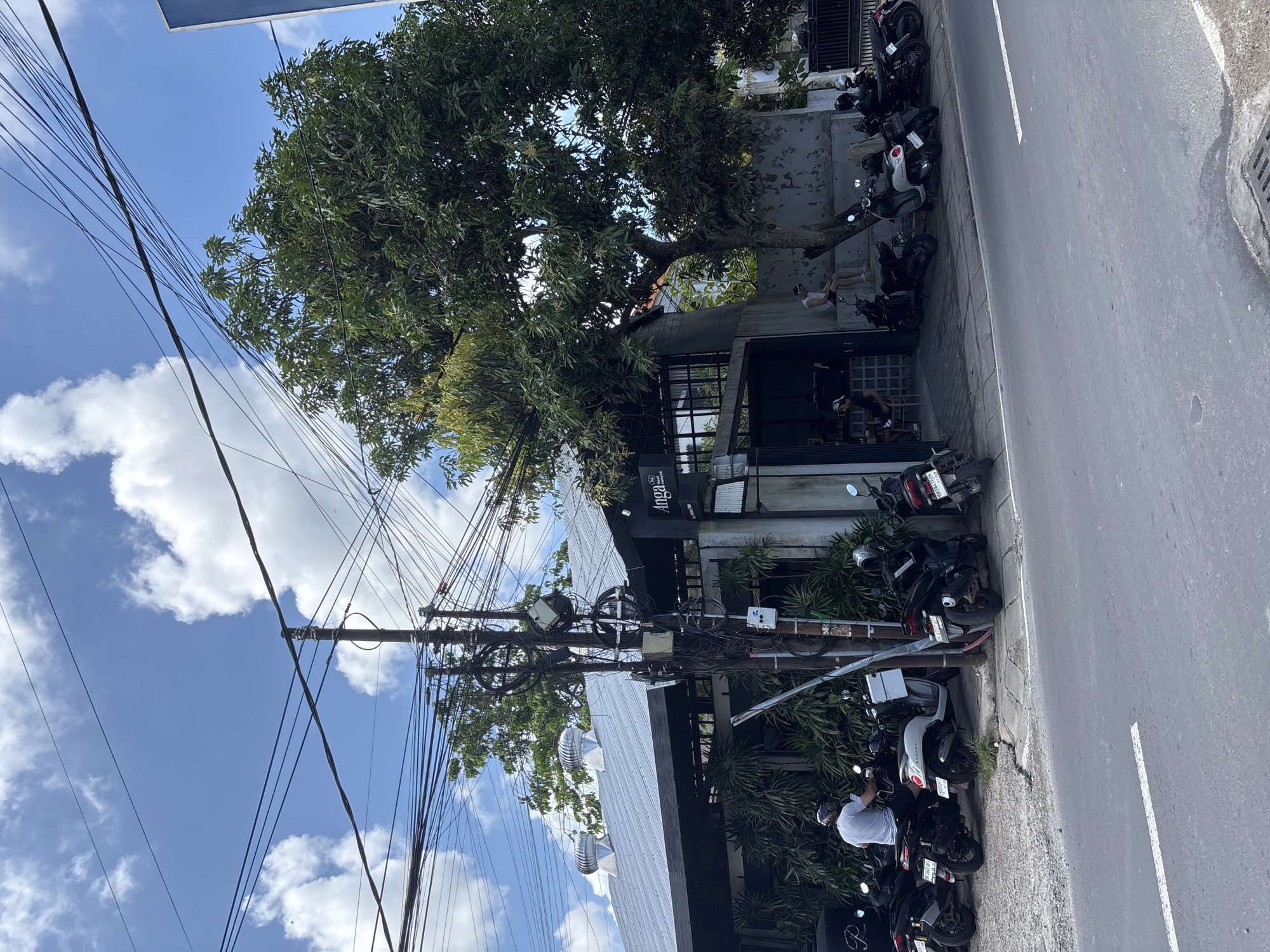 The image captures a street scene in Kuta Selatan, Indonesia, showing a street corner with a two-story building housing a business named "Anga". The building is light-colored with a black-framed window area. A large tree overhangs the structure, its branches extending toward the street. Multiple motorcycles are parked along the curb in front of the building. Utility poles are visible with a dense network of overhead wires spanning across the sky. The sky is bright blue, dotted with scattered white clouds. The street surface is gray asphalt with a white dividing line. Patrons are visible sitting in the window area of the "Anga" building. A person in a white shirt is seen standing near the motorcycles.