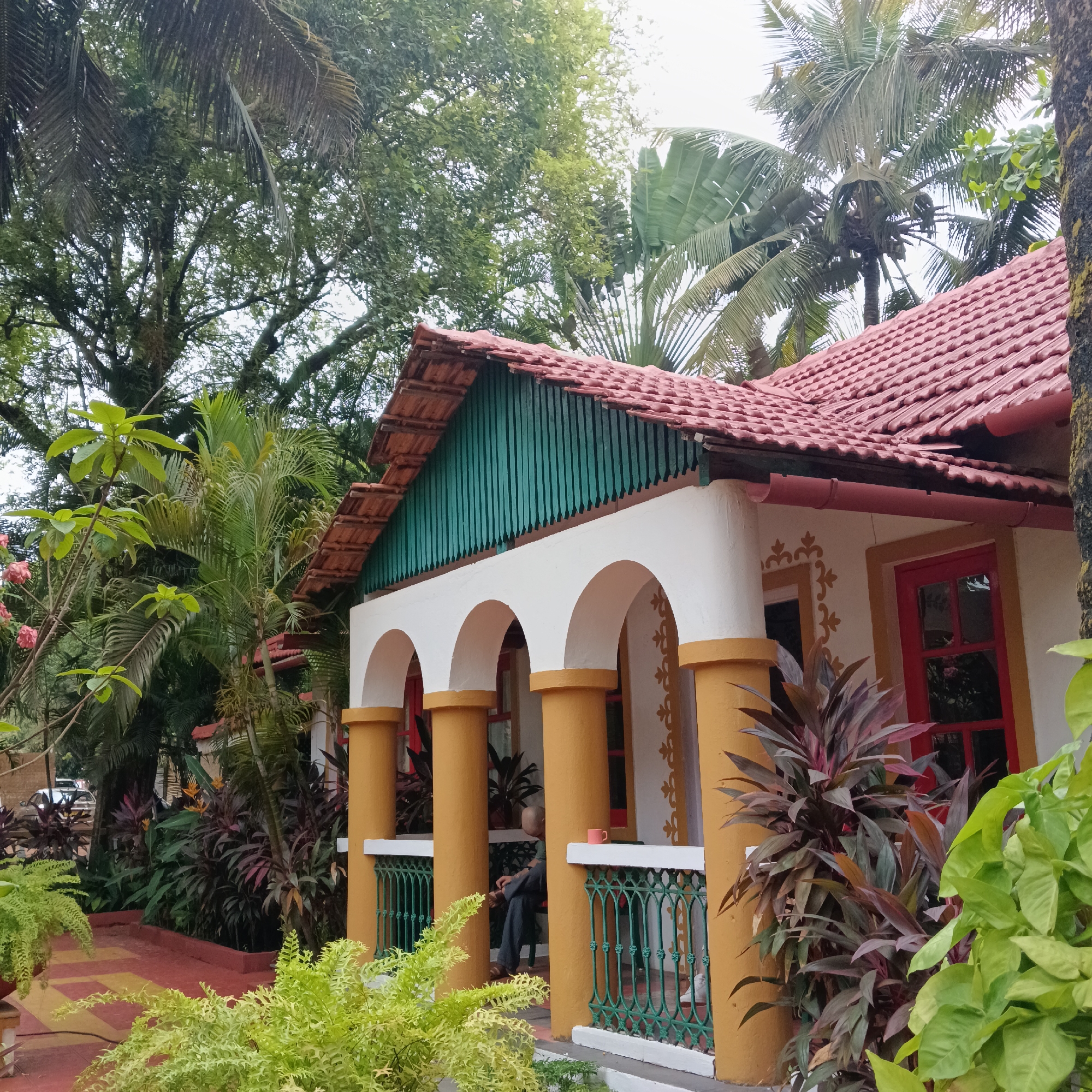A low-angle shot of a tropical-style building in Anjuna, India, featuring cream-colored walls, orange trim, and a red tile roof, surrounded by lush vegetation. The building's facade includes a veranda with orange cylindrical columns supporting white arches. A green vertical-planked attic area sits beneath the roof. A man sits on the veranda. Green wrought-iron railing is visible. A patterned red and yellow brick path leads to the building, and various tropical plants and trees, including palms, fill the foreground and background. A red-framed window and ornate detailing on the walls provide further visual interest. The scene is set during the day under overcast skies.