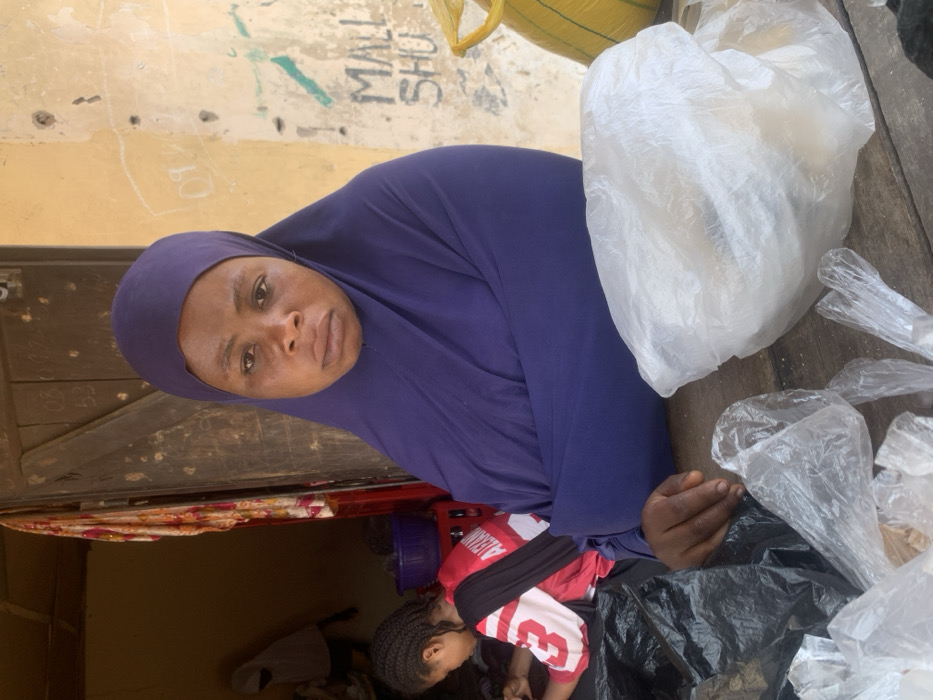 The image depicts an outdoor market or shop entrance in Okene, Nigeria. In the foreground, an adult female with a neutral expression, wearing a dark purple hijab, looks directly forward from behind a weathered wooden counter. Her right hand rests on the counter. Displayed on the counter are multiple clear plastic bags, one containing light-colored goods, and a black plastic bag. To the left, a younger female with braided hair, seen from the back, wears a red and white shirt displaying 'ALEXANDR' and is engaged in an activity within the dimly lit interior. Another person in a light hijab is partially visible further inside. A weathered wooden door, marked with numbers (e.g., '08', '0312', '53'), frames this interior. To the right, a stained, light yellow exterior wall features green graffiti spelling 'MALL SHU' and other faint markings. A yellow sack is partially visible on the far right.