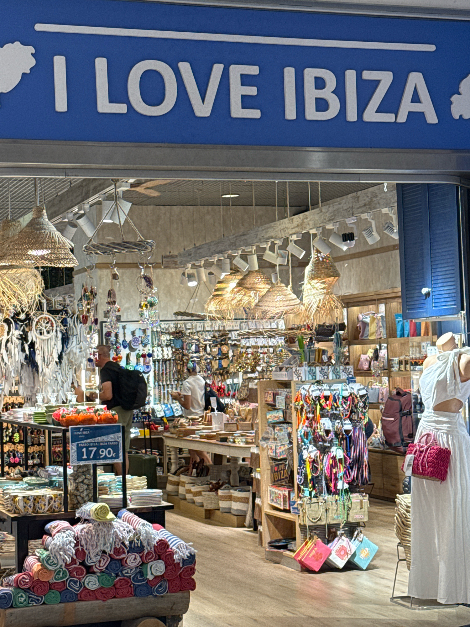 The image shows the exterior and interior of a shop with a blue awning displaying "I LOVE IBIZA" in white letters. The shop is filled with a variety of merchandise, including colorful textiles, dreamcatchers, jewelry, bags, and decorative items. In the foreground, a pile of rolled-up towels in various colors is displayed. Inside the shop, several shelves are stocked with goods. Two men are visible inside the shop, one wearing a black shirt and the other a white shirt. A mannequin dressed in a white dress stands near the entrance, holding a pink handbag. The shop appears to be located in a tourist area, possibly in the city of Sant Josep de sa Talaia, Spain, given the "I Love Ibiza" signage. The overall atmosphere is that of a vibrant souvenir or boutique shop.
