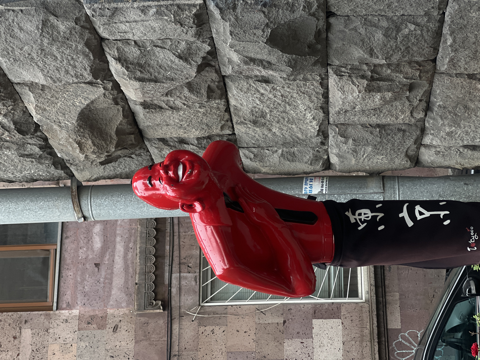 The image, taken in Yerevan, Armenia, shows a red sculpture of a face wearing boxing gloves. The sculpture is attached to a metal pipe running horizontally along the exterior of a building. The building's facade is constructed of gray stone blocks. The person wearing the boxing gloves is wearing black clothing with white lettering. A window with a metal grate, a partial view of a car, and a graffiti-like design on the building's wall are visible in the background.