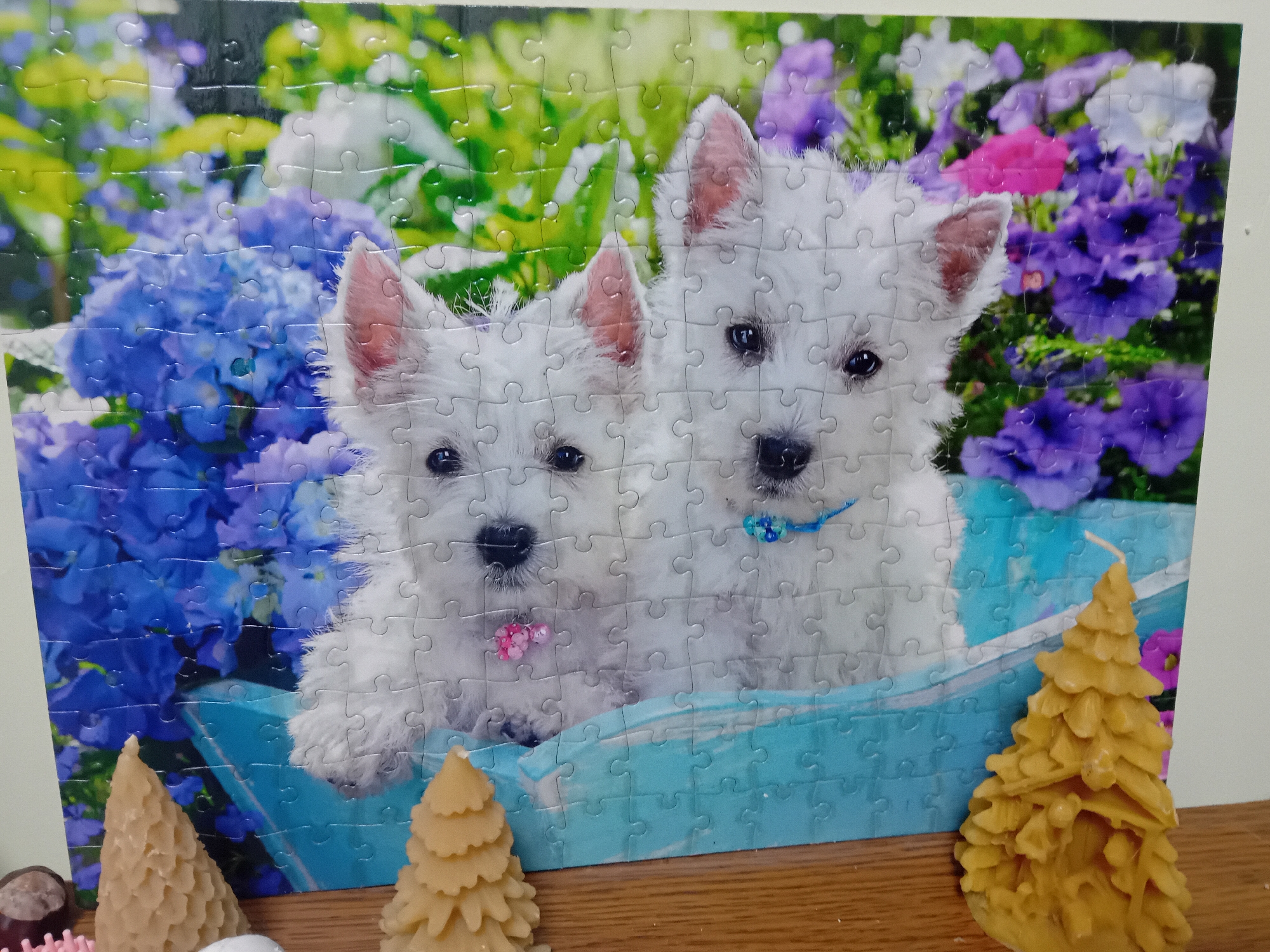 The image shows a close-up of a completed jigsaw puzzle depicting two West Highland White Terrier puppies sitting in a blue basket surrounded by blue and purple flowers. In the foreground, on a wooden surface, are three beeswax candles shaped like Christmas trees. The puzzle is leaning against a plain wall. The location of this scene in Chuhuiv, Ukraine is not discernible from the image.