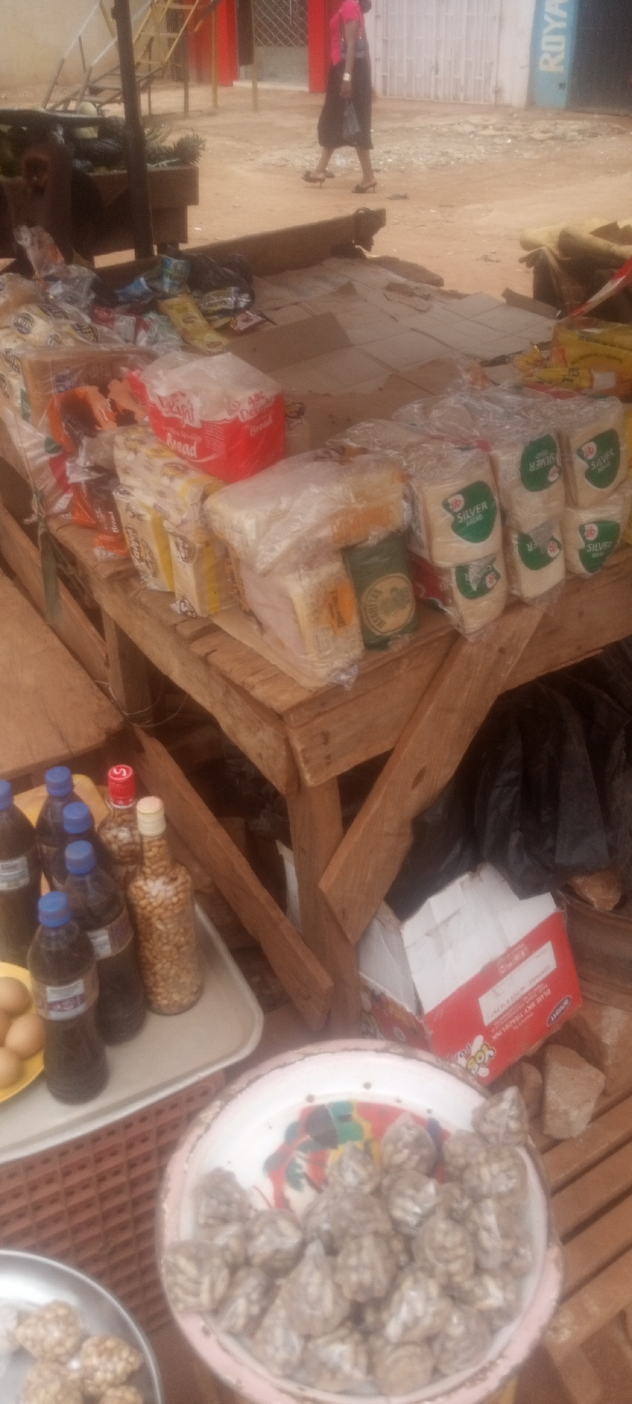 The image depicts a street-side vendor's stall in Sabon Tasha, Nigeria. The stall, constructed of wooden planks, displays a variety of goods for sale. Several loaves of sliced bread, some packaged with the brands "ABC Delight Bread" and "Silver Bread," are prominently featured. A collection of glass bottles, some filled with peanuts and others with a dark liquid, sit on a tray alongside a plate of eggs. In the foreground, a shallow metal dish overflows with small, individually wrapped items. A red and white cardboard box is visible beneath the table. In the background, a person wearing pink is walking away from the stall. The surrounding environment suggests an outdoor, potentially dusty, market setting. A building with a blue sign reading "ROYA" can be seen partially.