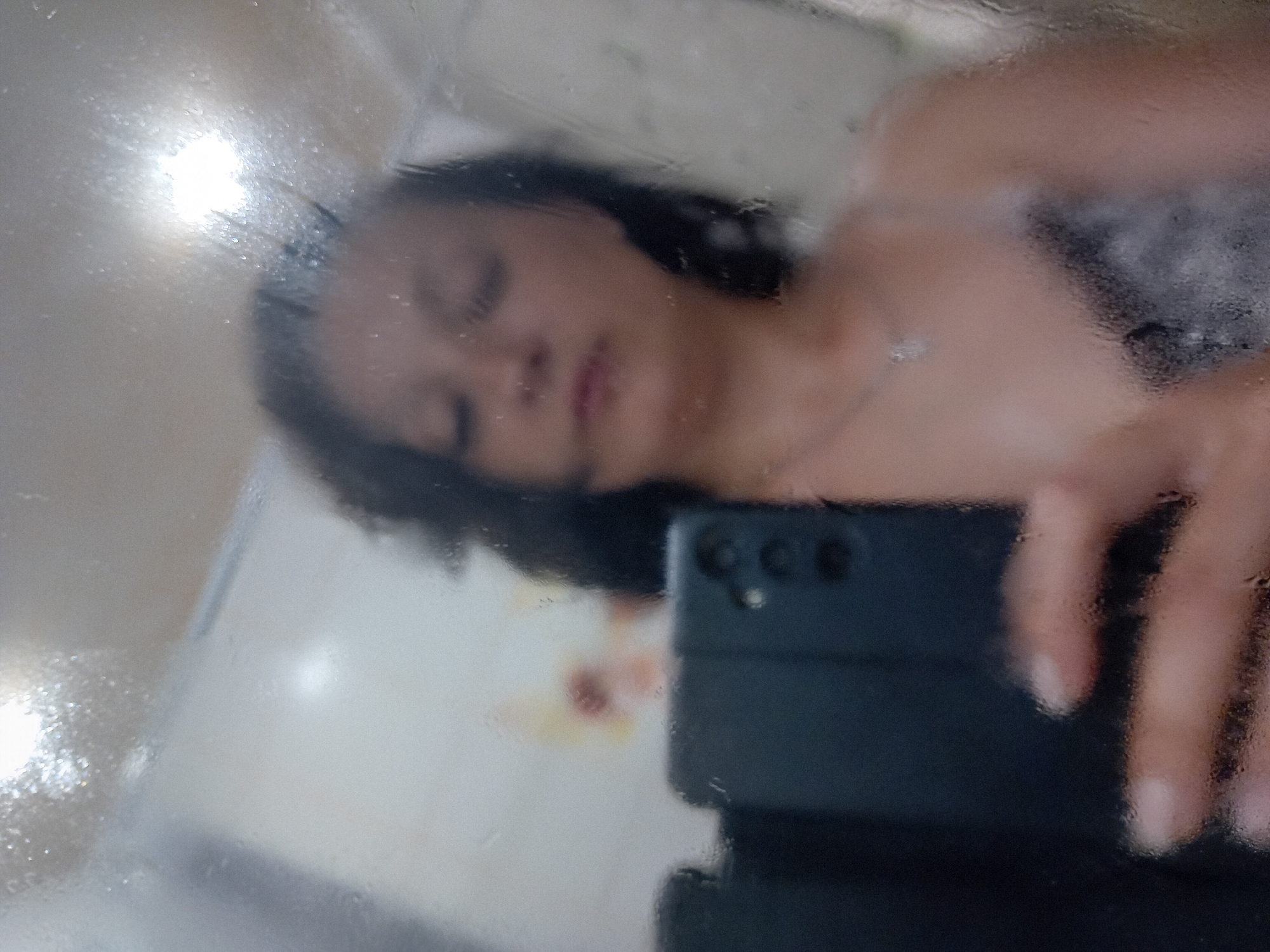 A selfie taken in a bathroom is reflected in a mirror covered with condensation. A woman with dark hair, wearing a light-colored top with black trim, holds up a smartphone to take the picture. Her face is partially visible, with her eyes closed and her lips slightly parted. The background of the bathroom is blurred, but a light fixture and a tiled wall are discernible. The image is framed by the condensation on the mirror, which creates a hazy effect. The location is specified as Chuhuiv, Ukraine.