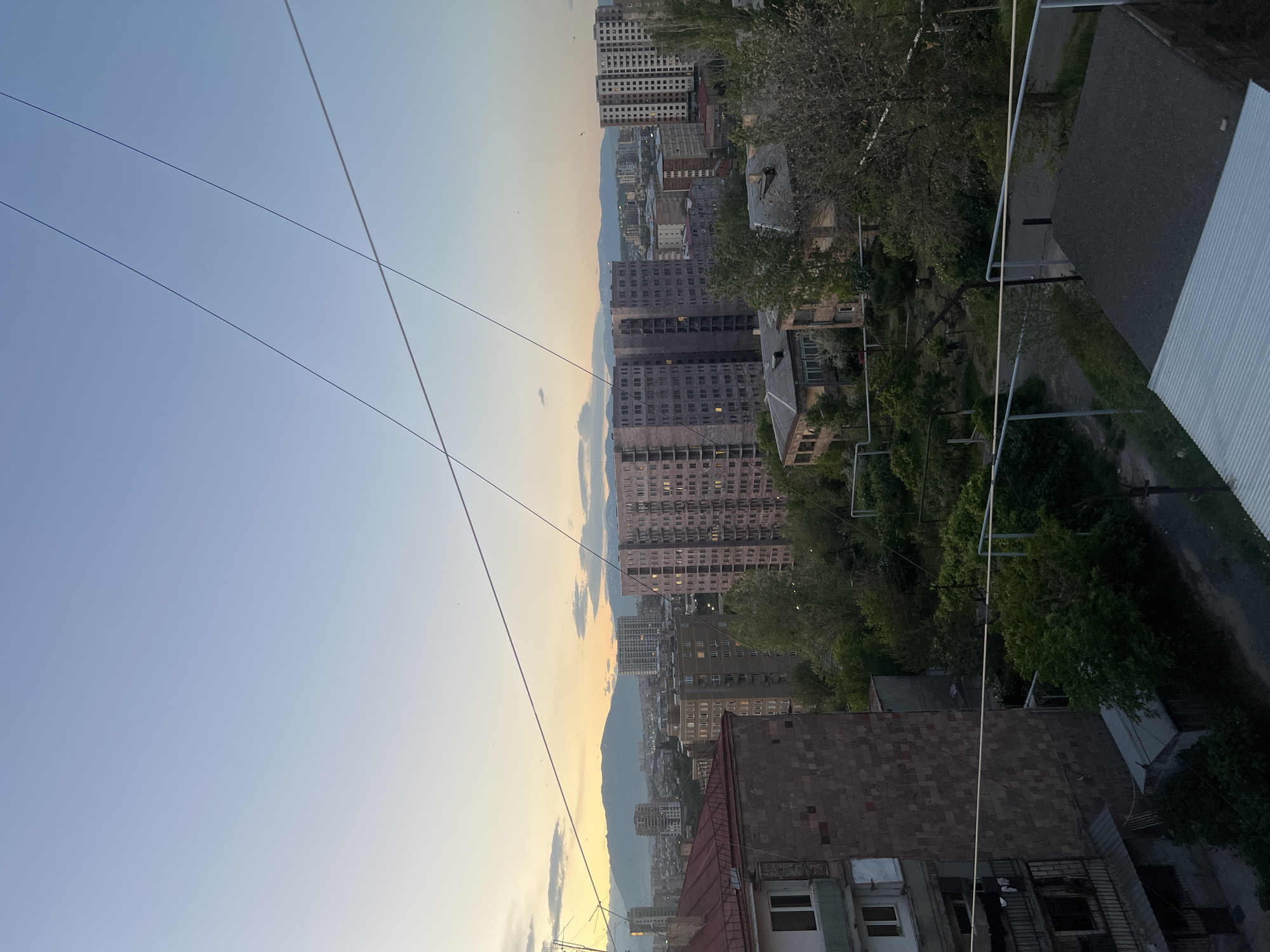 This image depicts a view from a high vantage point, likely a balcony or rooftop, overlooking a cityscape. The scene includes a mix of old and newer residential buildings. In the distance, there is a mountainous region, suggesting the city is situated in a valley or near a range. Overhead power lines are visible in the foreground, crisscrossing the sky. The sky is light blue, indicating either early morning or late afternoon. Based on the architectural style and mountainous background, this is likely Yerevan, Armenia.