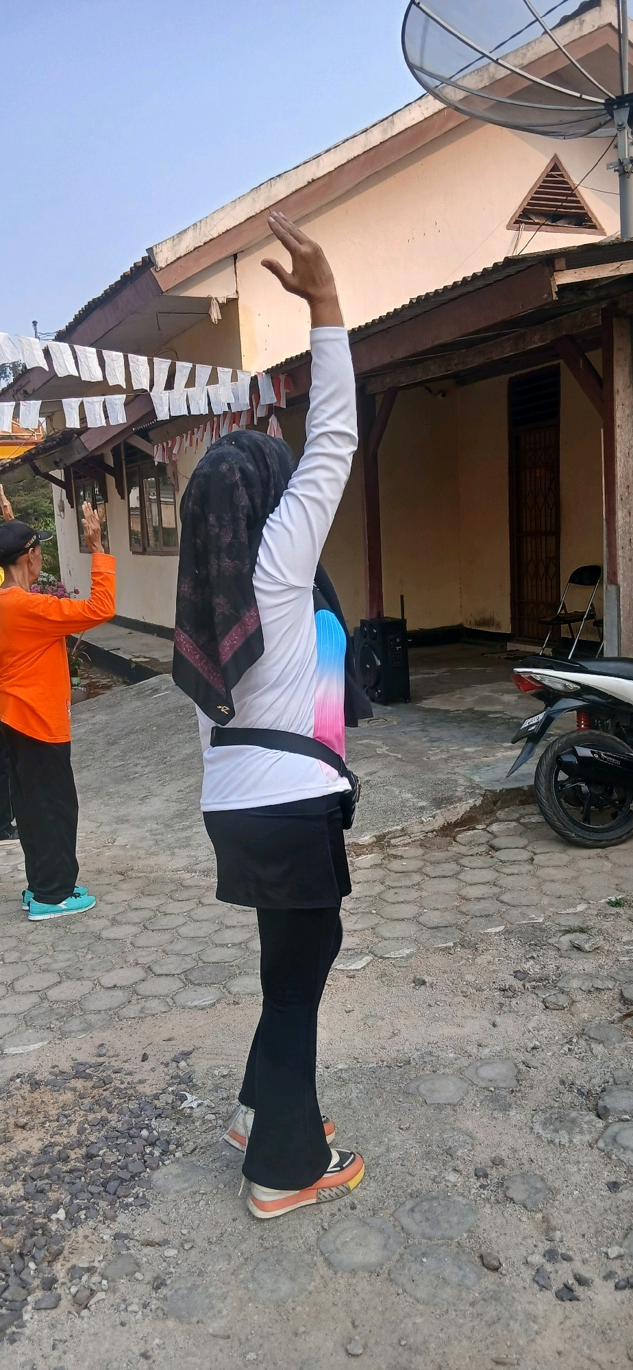 The image shows a side view of two individuals outdoors, presumably in the city of Metro, Indonesia. The person in the foreground is a woman wearing a black hijab, a white long-sleeved shirt with a gradient of blue and pink on the sleeves, a black skirt over black leggings, and colorful sneakers. She has her right arm raised in the air, palm open. Behind her, to the left, is another person wearing an orange long-sleeved shirt and black pants, with their arms also raised. The ground is paved with hexagonal stones and dirt. In the background, there is a building with a brown roof, an antenna, and decorative white and red flags strung across the top. A motorcycle is parked to the right of the frame. The overall scene suggests an outdoor activity, possibly exercise or a gathering, taking place in what appears to be a residential or community area.