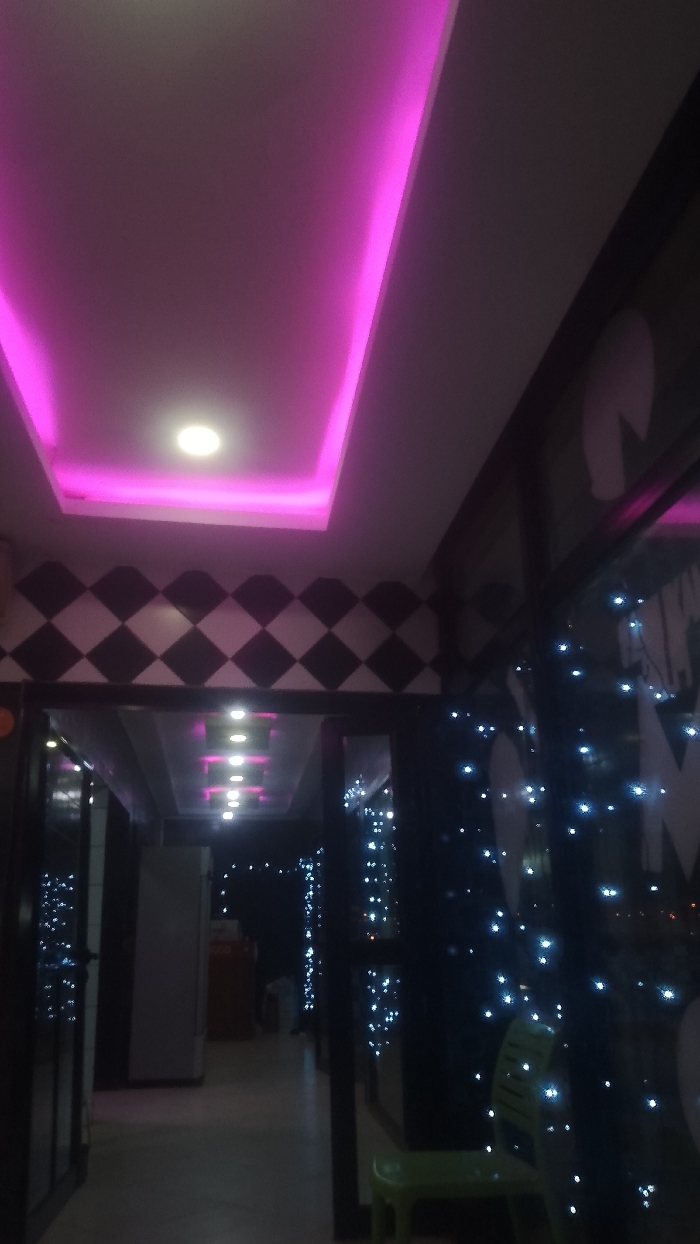 A hallway is illuminated by pink neon ceiling lights and recessed fixtures. Above the doorway, a checkered pattern borders the ceiling. Further down the hall, lights mimic the overhead design. To the right, sparkling lights decorate a window or divider, obscuring the view beyond. A green plastic chair sits in the foreground near the decorative lights. The overall impression is of a festive or themed interior space.