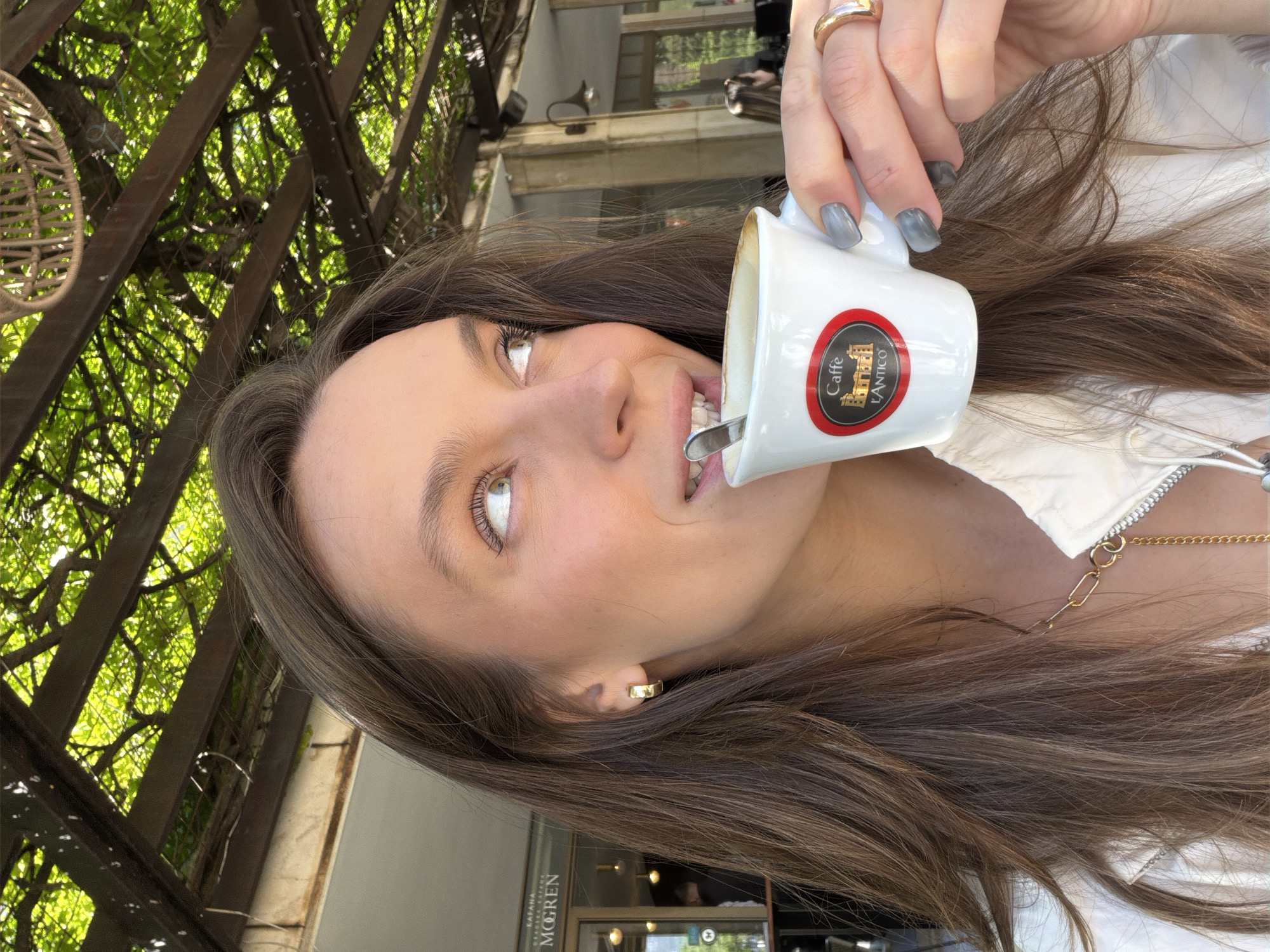 A candid, brightly lit portrait captures a young woman enjoying a cup of coffee in an outdoor setting, likely a cafe or restaurant patio in Budva, Montenegro. The image is taken at an angle, showing the woman looking upwards with a relaxed smile, revealing her upper teeth. Her long, dark brown hair cascades around her shoulders and chest. She wears a white collared shirt or jacket and accessorizes with a delicate gold chain necklace, a small gold hoop earring, and a gold ring on her right index finger. Her fingernails are painted a light gray-blue.
She holds a small white espresso cup with a red circular logo that clearly reads "Caffè L'ANTICO" and features an illustration of an old building. A small metal spoon is visible inside the cup as she takes a sip.
The background is dominated by lush green foliage, possibly vines or ivy, growing on a wooden or metal pergola structure, suggesting a shaded outdoor area. Beyond the foliage, glimpses of building architecture are visible. Partially visible text on a building in the background appears to read "KAFANA MOGREN," which strongly indicates the location is near Mogren Beach in Budva, Montenegro, consistent with the provided context. The bright lighting suggests it is daytime. The overall impression is one of leisure and enjoyment in a pleasant, natural environment.