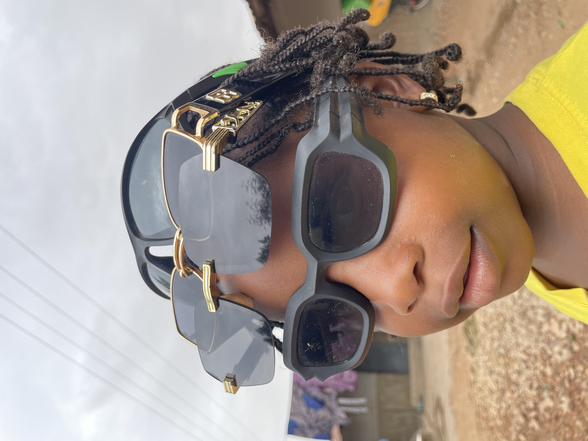 A person with braided hair is wearing two pairs of sunglasses stacked on their head. The top pair has a black frame with gold accents, and the bottom pair is matte black. A gold ring is visible in one of the braids. The person is wearing a yellow shirt, and the background shows an outdoor scene with a grey sky and blurred vegetation. Based on the request, the location is Jos, Nigeria, but there are no specific identifiers within the image to confirm this.