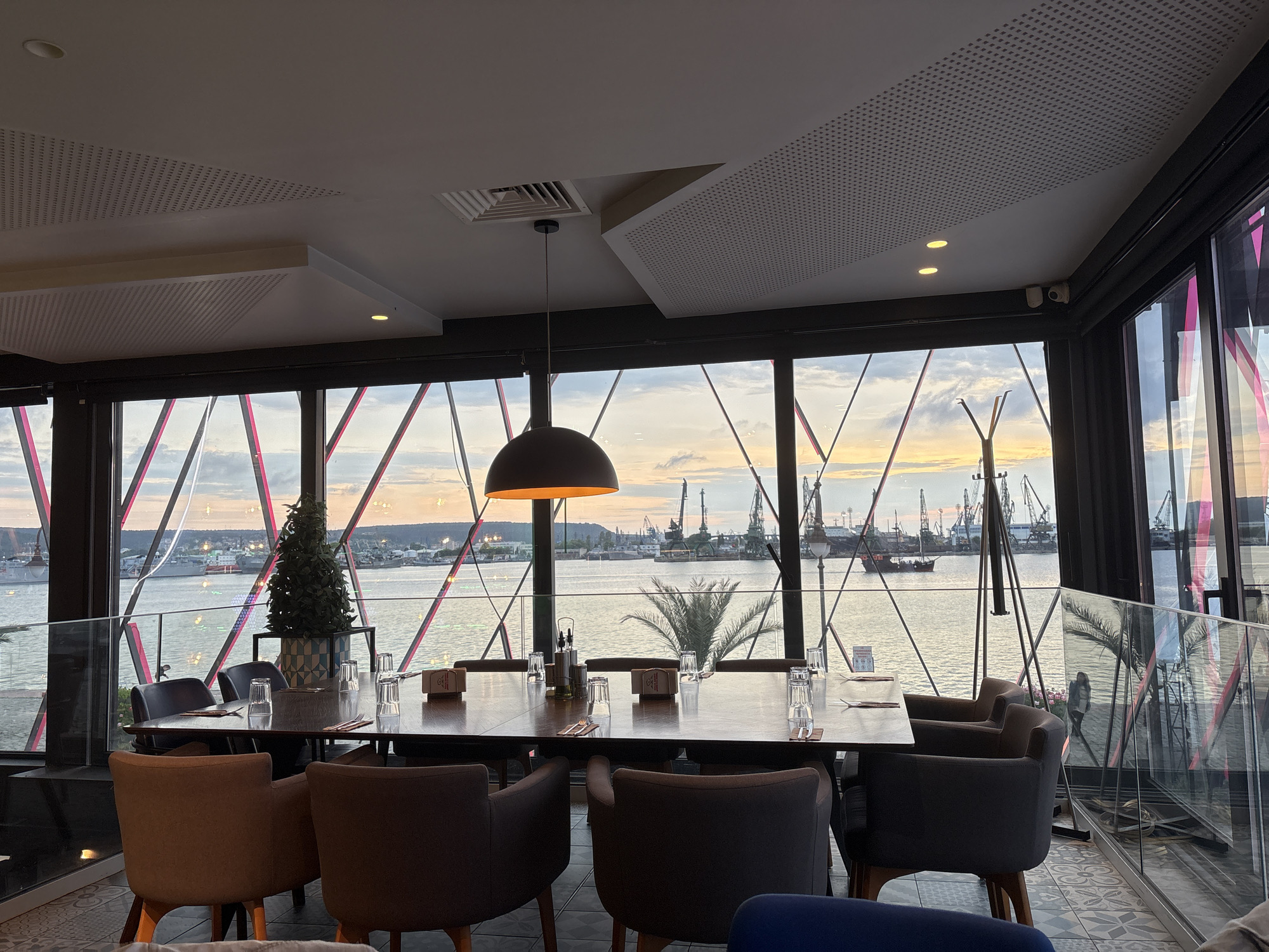 This media file presents a sophisticated interior space, likely a restaurant or cafe, offering a panoramic view of a harbor or port at either sunset or sunrise. In the foreground, a long, dark wooden dining table is neatly set with glasses and cutlery, awaiting guests. It is surrounded by several plush armchairs in dark gray and brown tones. A large, dark pendant lamp with a warm glowing interior hangs prominently above the table, providing ambient light.
The modern ceiling features white panels, some with perforated acoustic textures, interspersed with recessed spotlights and air vents. The space is defined by extensive floor-to-ceiling windows, characterized by a striking black framework that creates an intricate geometric grid of diagonal lines.
Beyond these windows, a large body of water reflects the soft, colorful hues of the sky—pinks, oranges, and blues—indicating the early morning or late evening. The view showcases an industrial port with numerous towering cranes lining the distant shore, suggesting active shipping operations. Several vessels, including a large cargo ship and a smaller, older-style boat with masts, are visible on the water.
Decorative elements inside include a small, conical artificial plant near the window and a natural palm frond visible through the glass. A glass railing partition is present on the right, and a faint reflection of a person can be seen within it. A small security camera is mounted near the top right window frame. Small logos or text on items on the table and a sign on the railing are present but unreadable. The scene evokes a calm, elegant atmosphere, contrasting the industrial view with the refined interior.