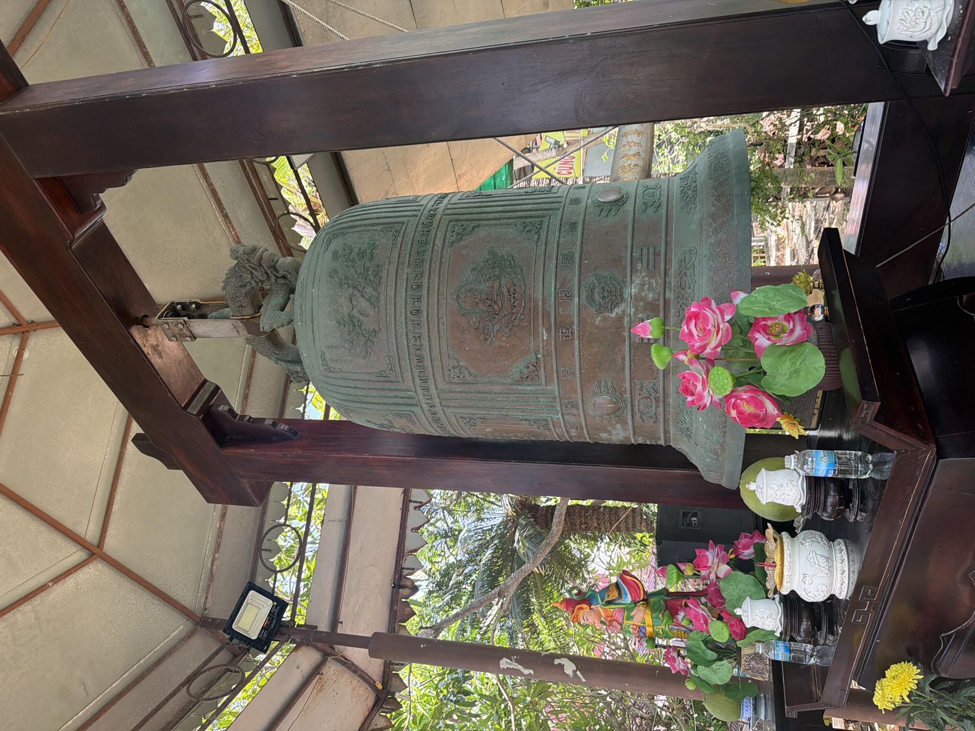 In Bac Nha Trang Ward, Vietnam, an oversized bronze bell, weathered to a greenish patina, is the central subject of this image. It hangs from a dark wooden beam structure, supported by thick, dark wooden posts. A small, intricately carved lion-like creature adorns the top of the bell. The bell itself is adorned with embossed relief carvings, featuring scenes and what appear to be inscriptions in an East Asian script.
Below the bell, a dark wooden altar displays several offerings. These include decorative white ceramic containers with lids, a yellow and pink artificial flower arrangement, and two bottles of water. A small, colorful statue of a human figure in traditional attire stands to the left of the bell. The altar is placed within a covered structure with a light-colored, possibly metallic, roof supported by a metal frame, and it is illuminated by a modern floodlight mounted on a metal pole. Lush green foliage and palm trees are visible in the background, suggesting an outdoor or semi-outdoor setting. The overall atmosphere is serene and suggests a religious or spiritual location.