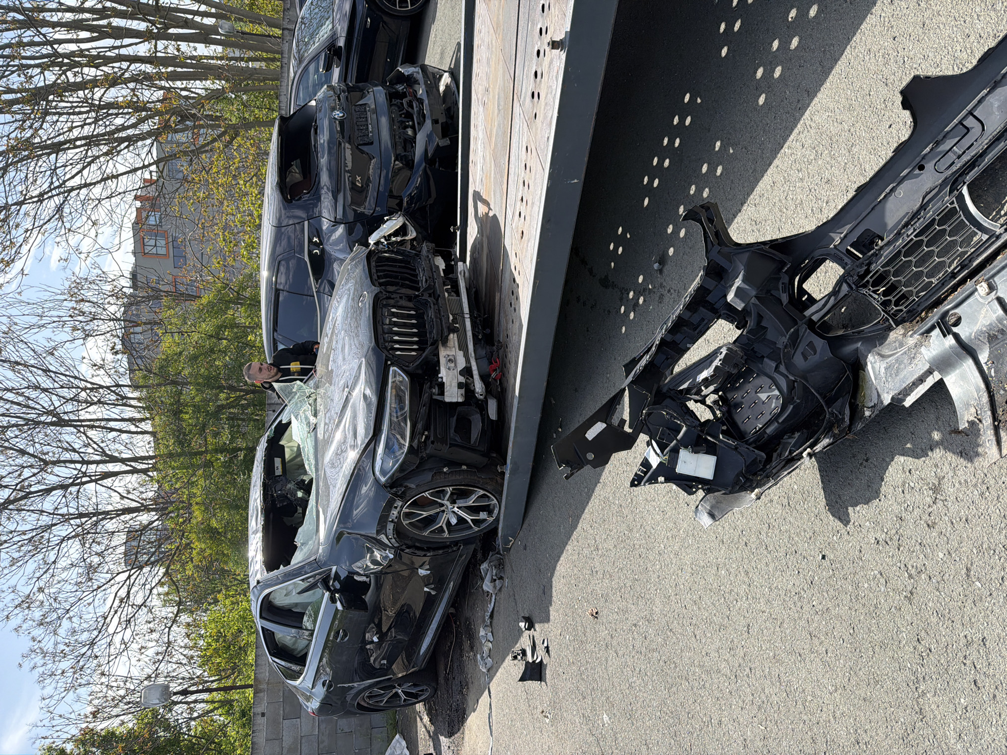 A wrecked black SUV is being transported on a flatbed tow truck. The front end of the SUV is severely damaged, with the bumper and other parts of the car lying on the ground nearby. The windshield is shattered, and the airbags have deployed. Another damaged black SUV is on the tow truck behind the first one. A man stands beside the first SUV, seemingly observing the scene. The incident appears to have taken place in an urban area, given the presence of buildings and trees in the background. The specific location, Burgas, Bulgaria, is indicated by visual markers in the image, notably architecture and landscape features. The scene suggests a vehicle accident.