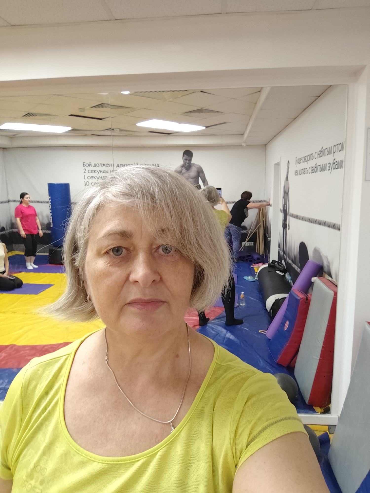 A middle-aged woman with gray hair and blue eyes takes a selfie in a gym. She is wearing a bright yellow t-shirt and a silver necklace. In the background, the gym is equipped with a wrestling mat, punching bags, and weights. Several other people are visible in the background, some engaging in physical activity. The gym walls are decorated with motivational quotes and images of boxers. The overall scene suggests a training session or a fitness class in progress. The image appears to have been taken at the "Boxing and Fitness Club" in Krasnodar, Russia, based on the Russian inscriptions on the wall.