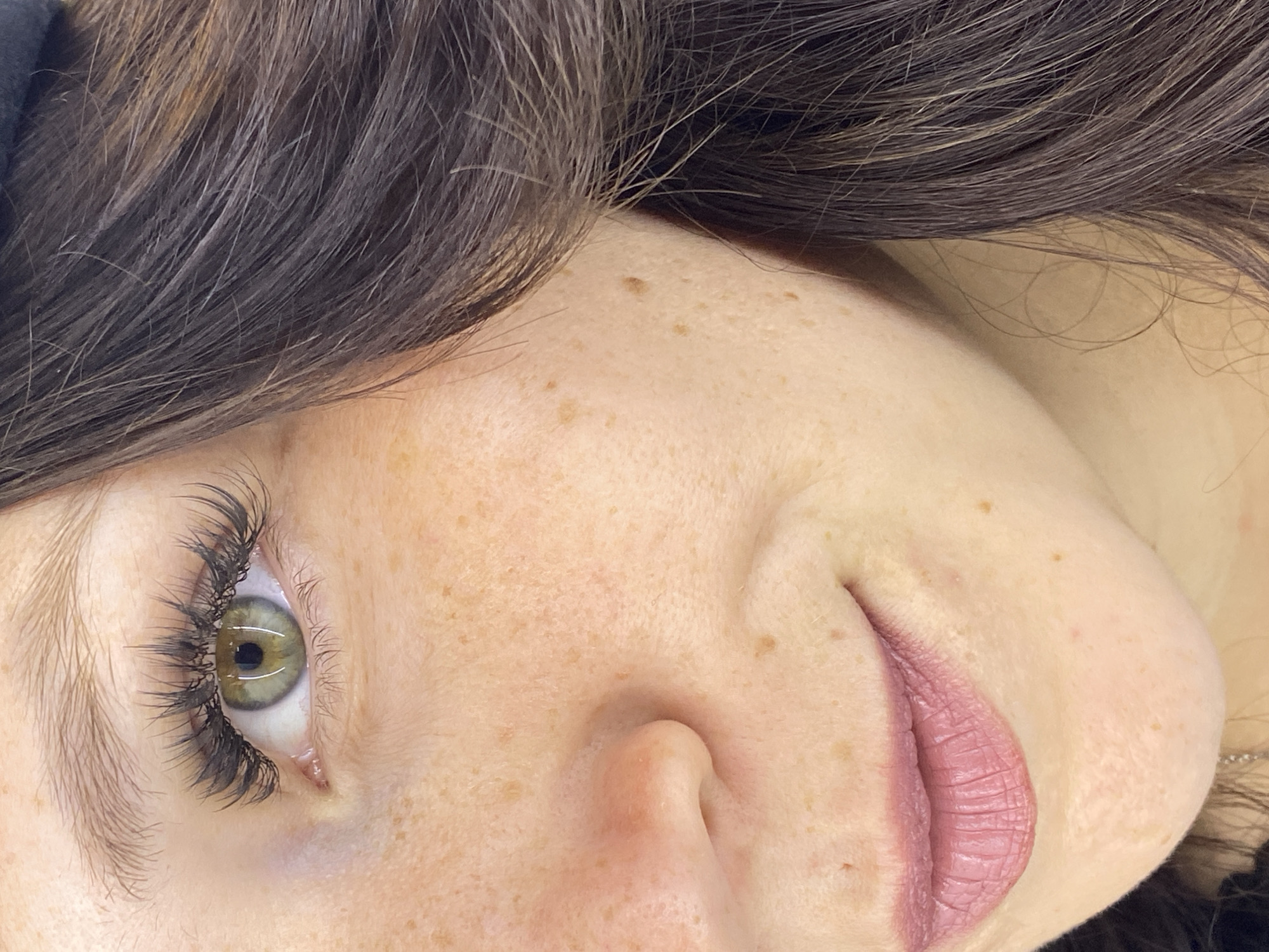 The image is a close-up of a woman's face, focusing on her left eye, nose, and mouth. She has green eyes with applied eyelash extensions and fair skin with freckles. Her lips are painted with a matte pink lipstick. Dark brown hair frames the right side of her face. There is no information about the location being in Chaikovsky, Russia.
