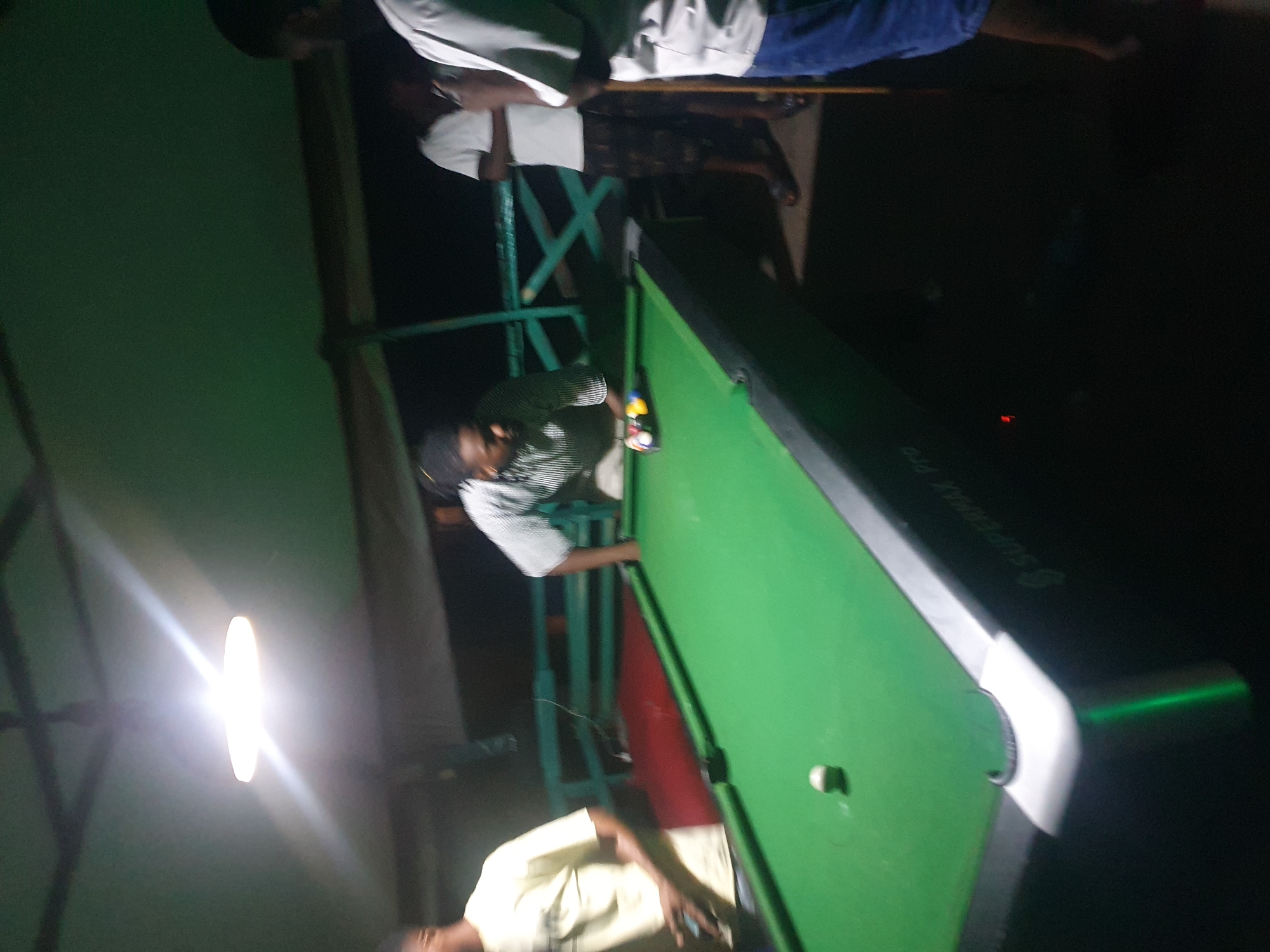 In Lokoja, Nigeria, a group of four men are gathered around a pool table. One man, wearing a striped shirt, is bent over the table, preparing to strike a billiard ball with a cue. Another man, in a yellow shirt, stands to the left, looking at the table. To the right, two other men are observing the game. The pool table is green, with several colored balls arranged in a rack. The scene is illuminated by a bright light fixture hanging from the ceiling, casting a warm glow over the area. The environment appears to be an outdoor or semi-outdoor setting, possibly a bar or recreational area, with a wooden railing visible in the background. The lighting is dim in the background, highlighting the players and the pool table.