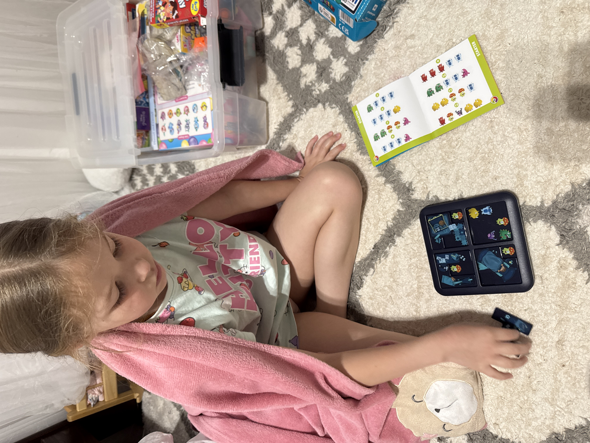 A young girl is playing with a puzzle on a light-colored rug with a gray pattern. She is sitting with her legs bent and covered with a pink blanket, wearing a light-colored pajama set with a Hello Kitty design. In her hand, she holds a puzzle piece. Next to her on the rug, there is a puzzle board and an open instruction booklet. To the left, there is a clear plastic storage bin filled with various items, including boxes and wrapped objects. The background includes a glimpse of a dark wood furniture piece and a white surface. The location information is not possible to determine from this image.