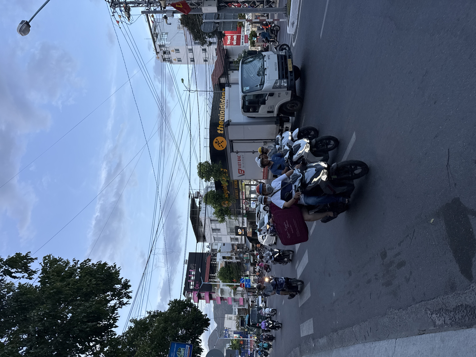 The image captures a bustling street scene in Bac Nha Trang Ward, Vietnam. A mix of scooters and motorbikes are stopped at a crosswalk, waiting for an opportunity to cross. A white delivery truck is parked on the side of the road near the crosswalk. The backdrop features buildings of varying heights and designs, and street signs. Utility lines crisscross the sky, adding to the layered and busy urban environment. The overall lighting suggests it is daytime with overcast or slightly cloudy conditions.