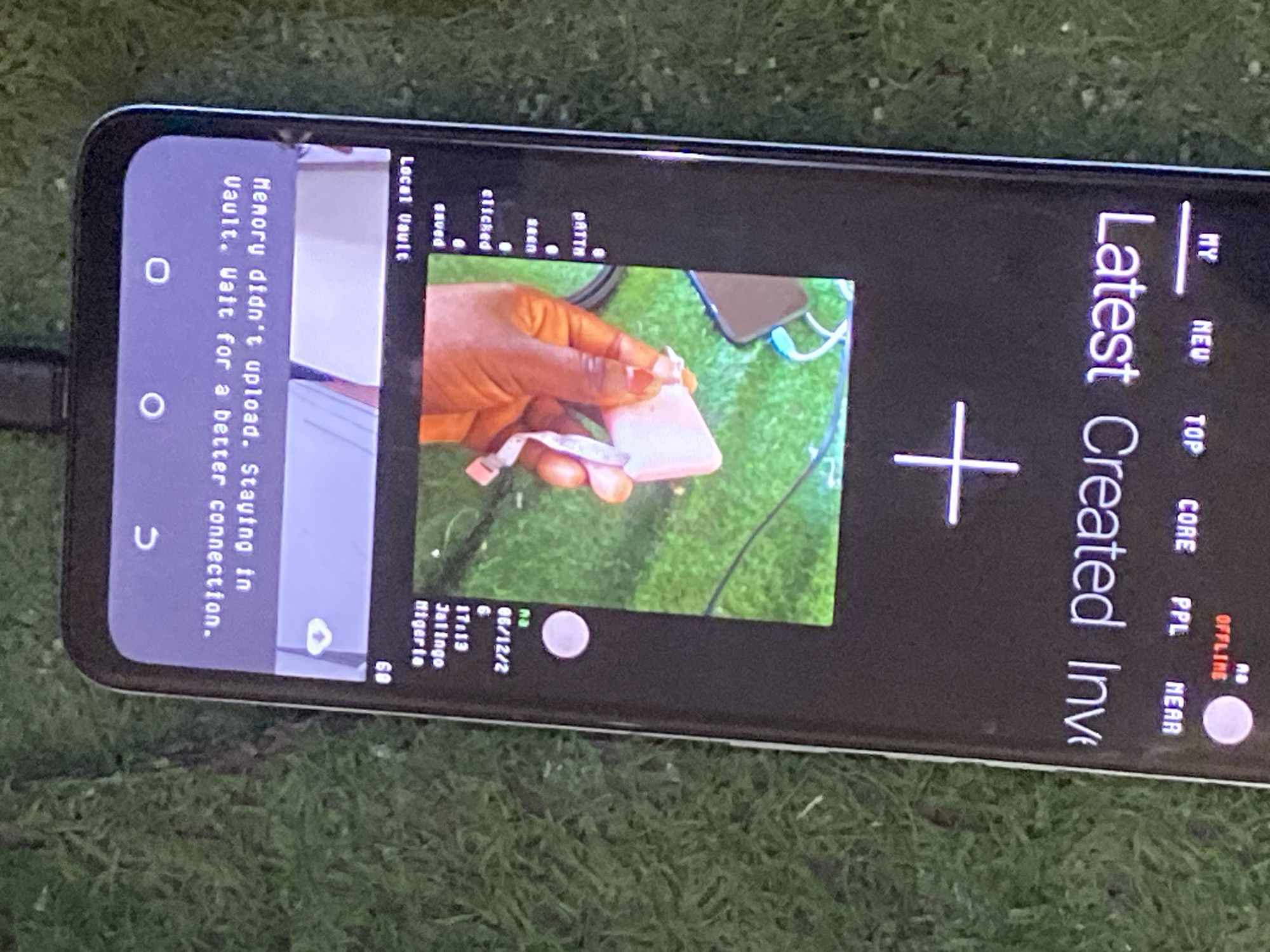 The image shows a smartphone lying on a grassy surface, possibly outdoors. The phone's screen displays a message "Memory didn't upload. Staying in vault. Wait for a better connection." The screen also shows a photo of a person's hand holding a small, pink object, with a blurry green background. There are also vertical text labels on the right side of the screen. One label reads "Latest Created Inv" and other smaller labels include "MY", "NEW", "TOP", "CORE", "PPL", and "OFFLINE". The location is identified as Jauro Yinu, Nigeria. The phone is angled and partially visible, with a black cable connected to its side. The grass in the background is green and appears somewhat dry. The lighting suggests it is daytime.