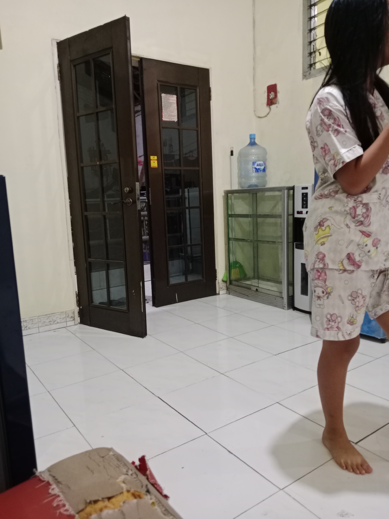 The image depicts an indoor scene, likely a residential area in West Jakarta, Indonesia. A young person, possibly a child or adolescent, is standing in the right side of the frame, wearing pajamas with a cartoon print. They are facing away from the camera, with their right leg visible and bare feet on a tiled floor.
In the background, a set of double doors, one ajar, stands to the left. These doors appear to be made of dark wood with glass panes. Further to the right, a large water dispenser bottle with an "AQUA" label is visible on top of a glass display cabinet. Adjacent to the cabinet is a white appliance, possibly a water cooler or a refrigerator. The floor is covered in white tiles with dark grout lines. A damaged piece of furniture, possibly a chair with exposed foam, is partially visible in the lower left corner of the image. The walls are painted a light cream color, and a red electrical outlet is mounted on the wall near the water dispenser. The lighting suggests an indoor environment with artificial light sources.