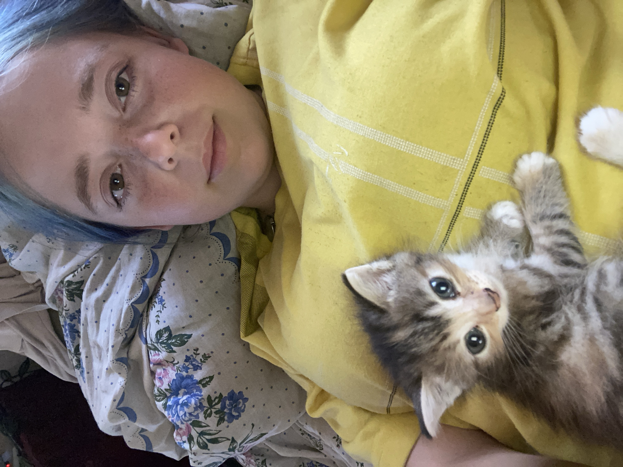 The image shows a young person with blue-dyed hair lying down, with a small, tabby kitten on their chest. The person's face is visible, and they are looking towards the camera with a gentle expression. The kitten is also looking at the camera, with large, dark eyes. Both subjects are resting on what appears to be a bed with floral patterned sheets and a yellow garment, possibly a sweatshirt or blanket. The scene suggests a moment of relaxation or quiet intimacy between the person and the kitten. The lighting appears to be natural, implying it could be daytime. There are no discernible signs of weather conditions or specific location cues beyond the general indoor setting. No text is visible in the image.