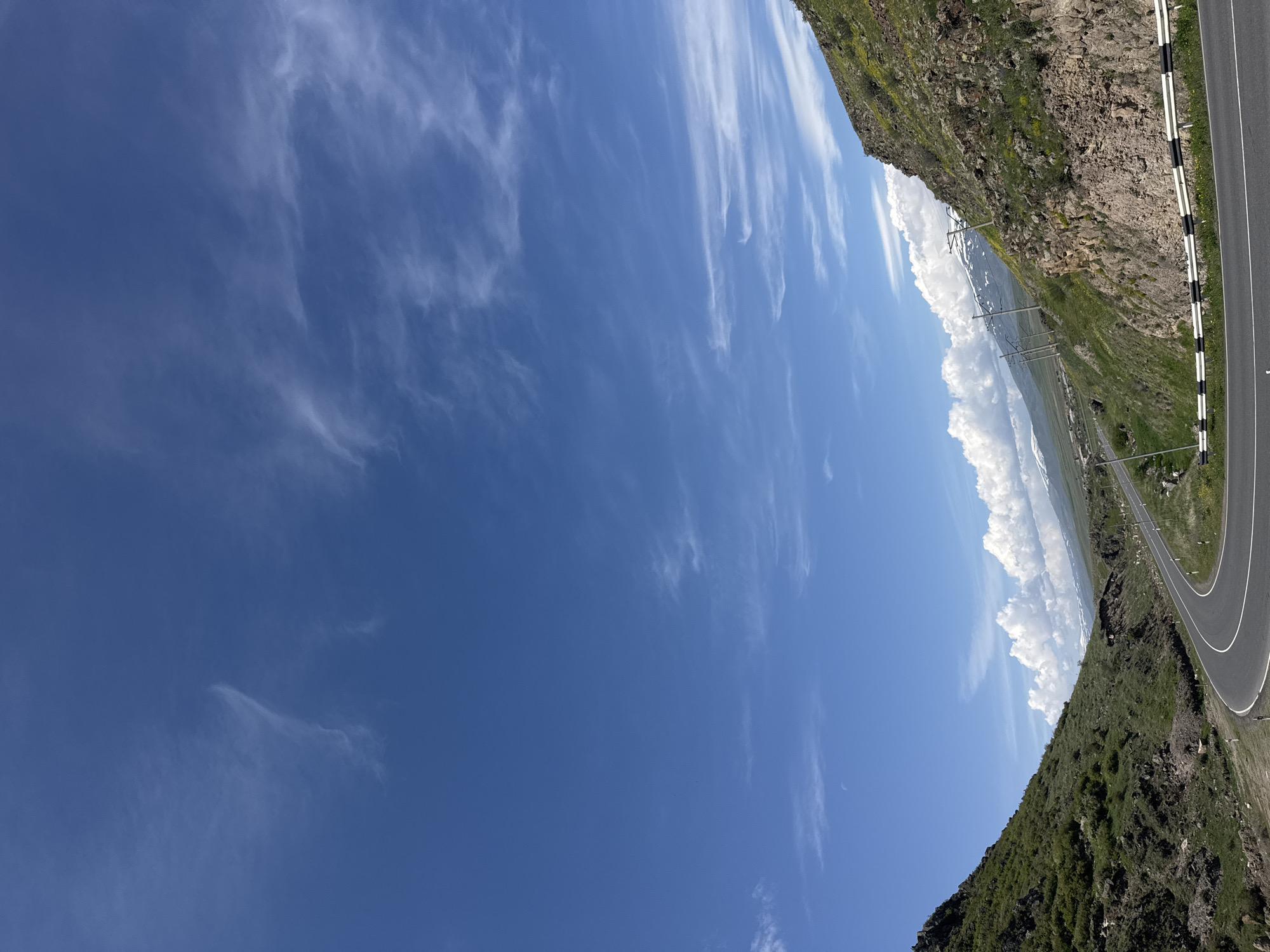 The image depicts a landscape near Bagravan, Armenia. A road with a black and white guardrail curves along the side of a rocky mountain covered with green vegetation. In the distance, there are white clouds and glimpses of snow-capped mountains. Power lines stretch across the landscape. The sky is predominantly blue with wispy, white clouds.