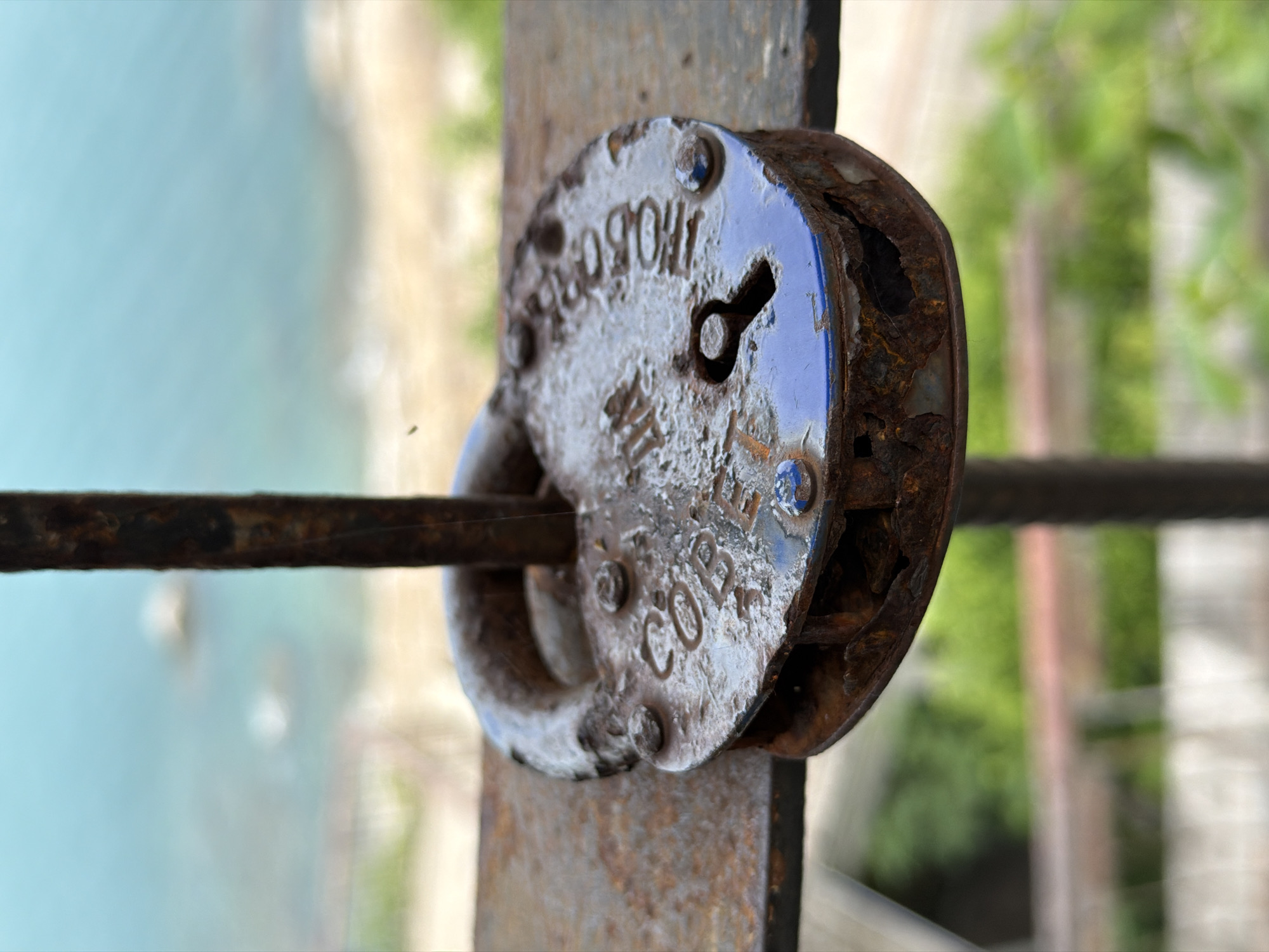 A close-up, high-angle shot focuses on a rusty, old padlock with blue accents, mounted on a metal railing. The padlock is oval-shaped and shows significant corrosion, with its metal casing visibly deteriorating in some areas, revealing darker, rusted material beneath. The words "COPPER" and "THOBO" are embossed on the surface of the padlock. A single, narrow, vertical metal bar, also rusted, passes through the shackle of the padlock.
The background is blurred, suggesting a shallow depth of field. In the upper portion of the image, a body of water, likely the ocean, is visible with a light blue hue. Distant, out-of-focus landmasses or large rocks can be seen in the water. Below the water, a sandy beach area and then out-of-focus green foliage are present, indicative of an outdoor setting. The overall scene suggests a coastal environment. The presence of "THOBO" which is a word commonly associated with the island of Koh Phangan, and the proximity to the sea strongly suggests the location as being along the coastline of Koh Samui District, Thailand, likely a resort or a scenic viewpoint overlooking the ocean. No people or specific actions are depicted.