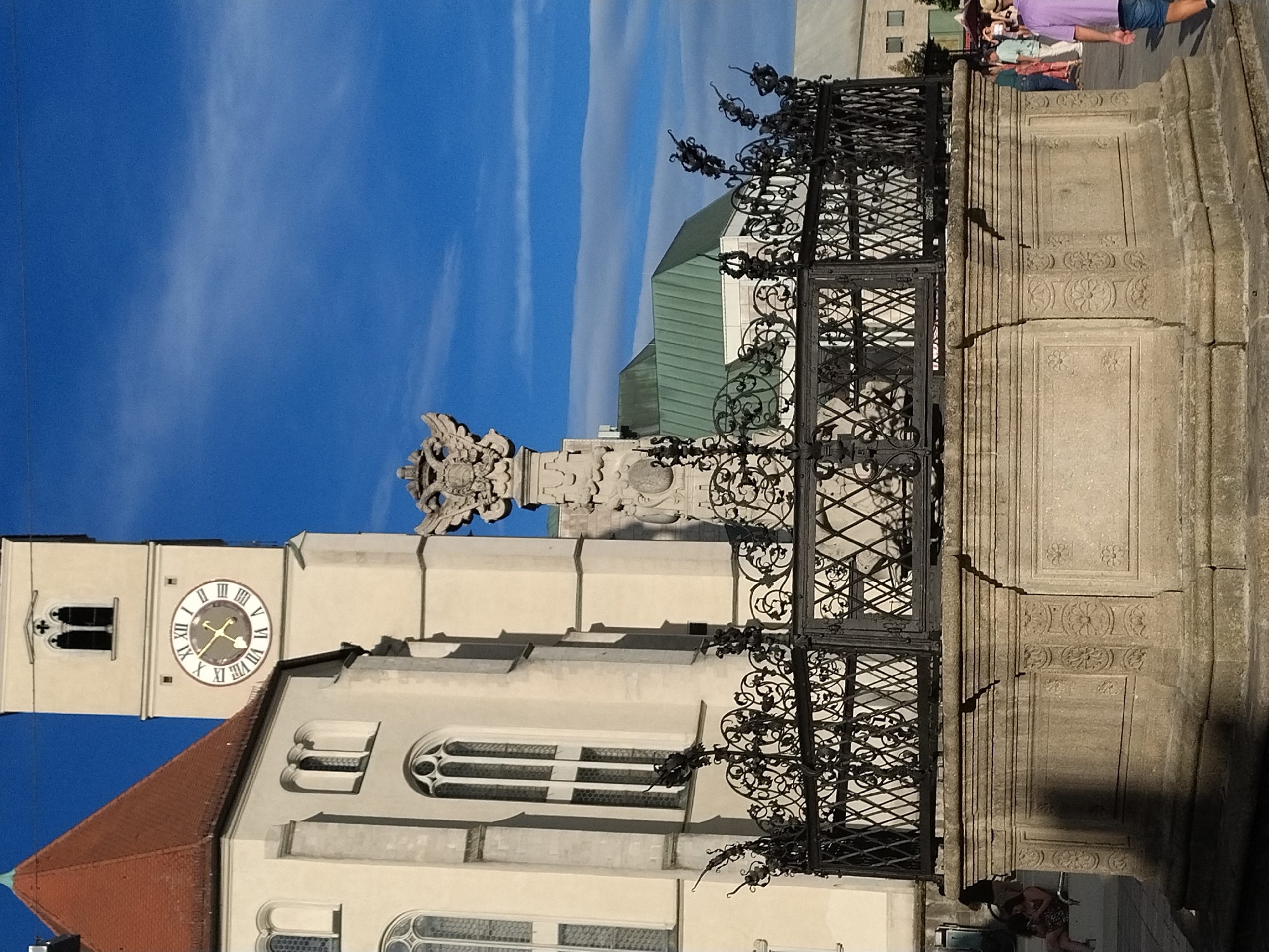 The image depicts a view of the St. Peter's Cathedral in Regensburg, Germany, specifically focusing on the historic Neptune Fountain. In the foreground, the stone base of the fountain is visible, adorned with decorative carvings of flowers and geometric patterns. A black wrought-iron fence surrounds the fountain, featuring intricate scrollwork. Above the fountain, a stone monument topped with a double-headed eagle emblem rises. To the left, a portion of the cream-colored facade of St. Peter's Cathedral is seen, with its distinctive red tiled roof and gothic-style windows. Adjacent to the cathedral is its bell tower, which has a clock face displaying Roman numerals. The sky above is a clear blue with some wispy clouds. In the background, other buildings and people are visible, suggesting a public square or marketplace. A person wearing a purple shirt and blue shorts is walking away from the camera on the right side of the image.