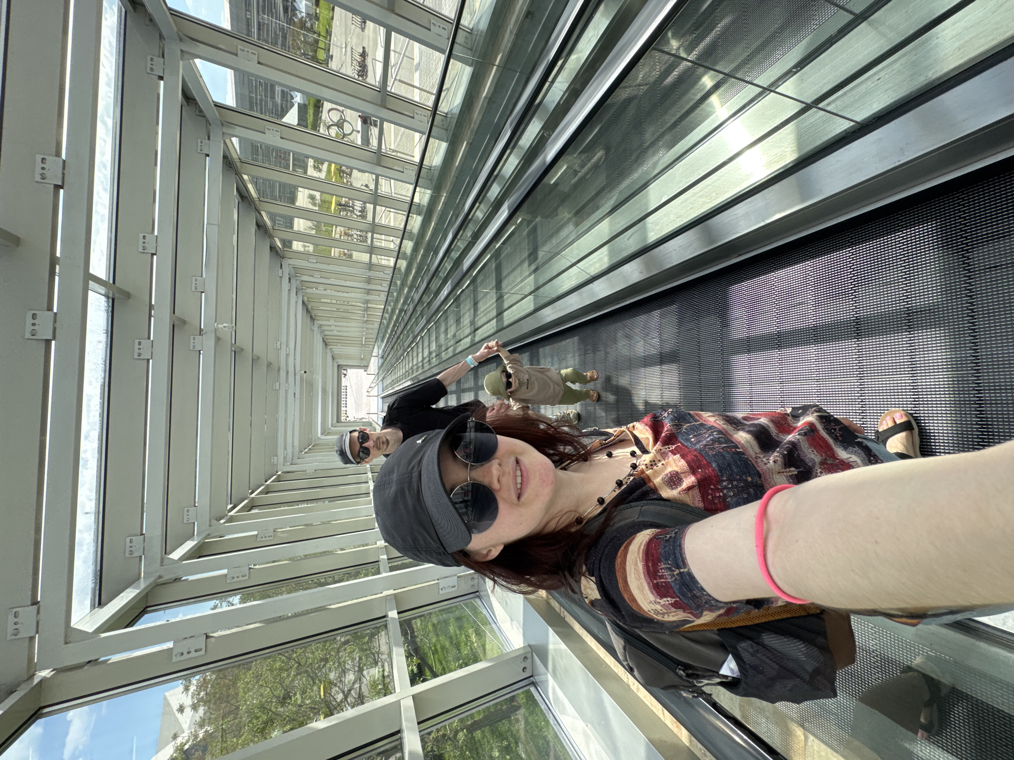 A selfie shows a woman on a moving walkway inside a covered structure. The structure is constructed with many windows and white metal framing. A man and a child are further down the walkway. The location is likely in the city of Sirius, Russia, based on the request.